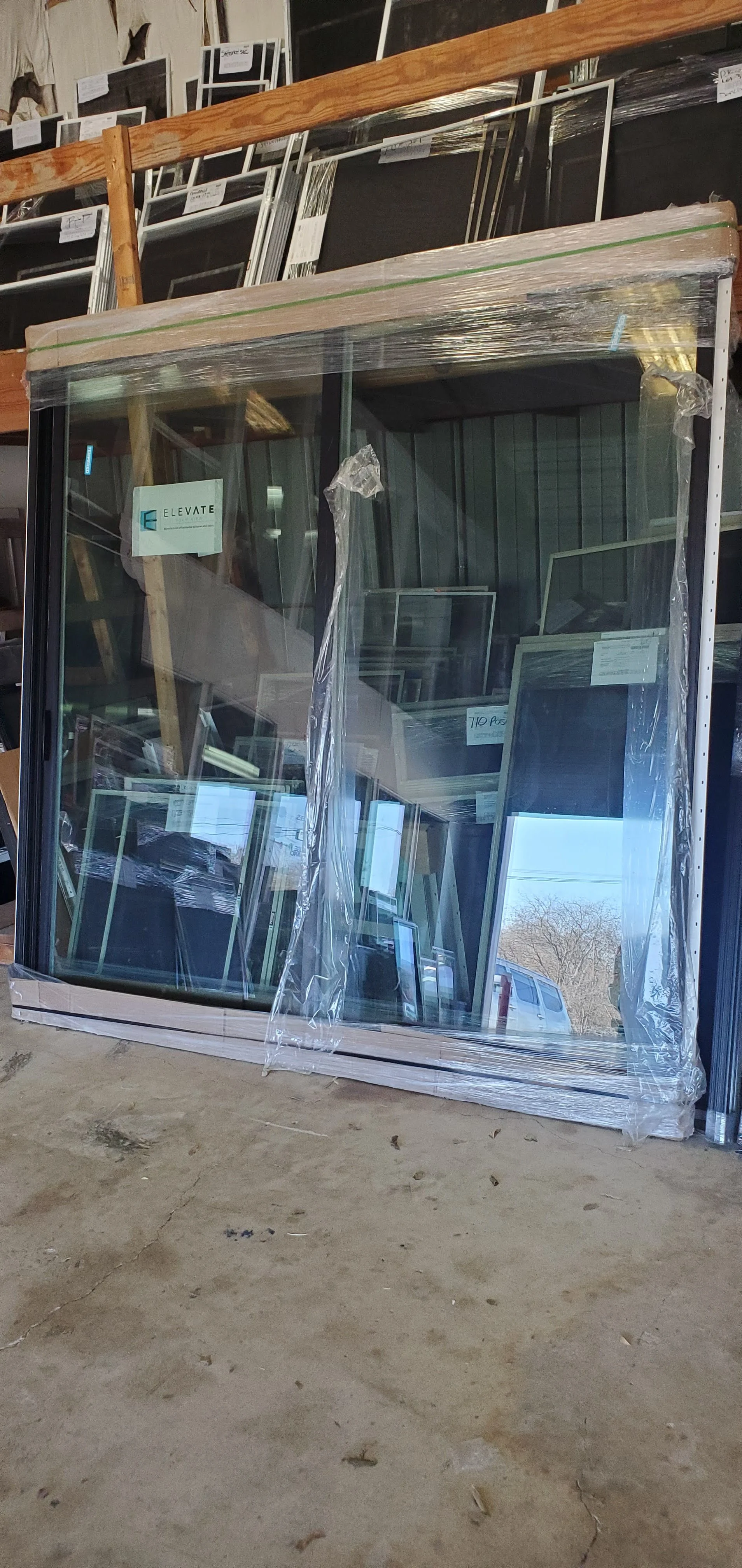 New window pane wrapped in plastic packaging leaning against a wall in a warehouse, with shelves of other glass window frames and a parking lot visible outside through the window.