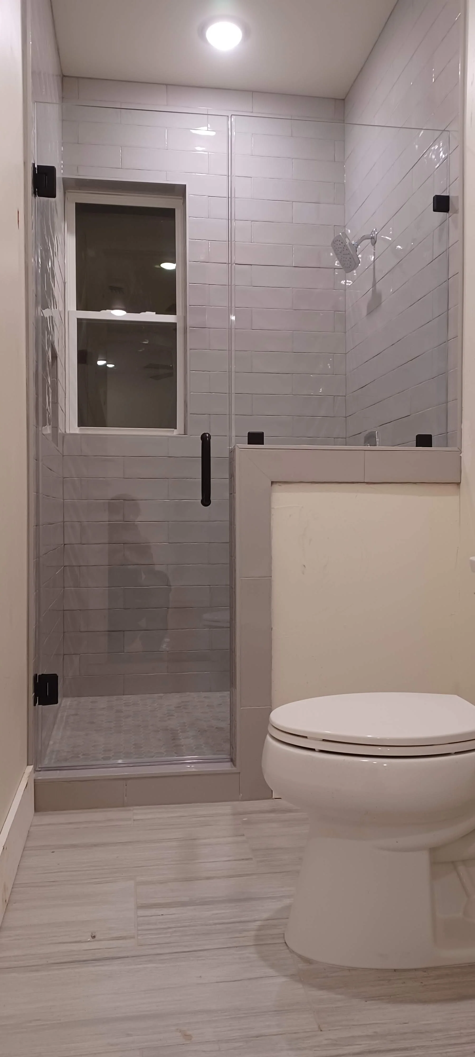 A bathroom with a glass-enclosed shower, a window, with white subway tiles on the walls, a ceiling light fixture, and a modern white toilet.