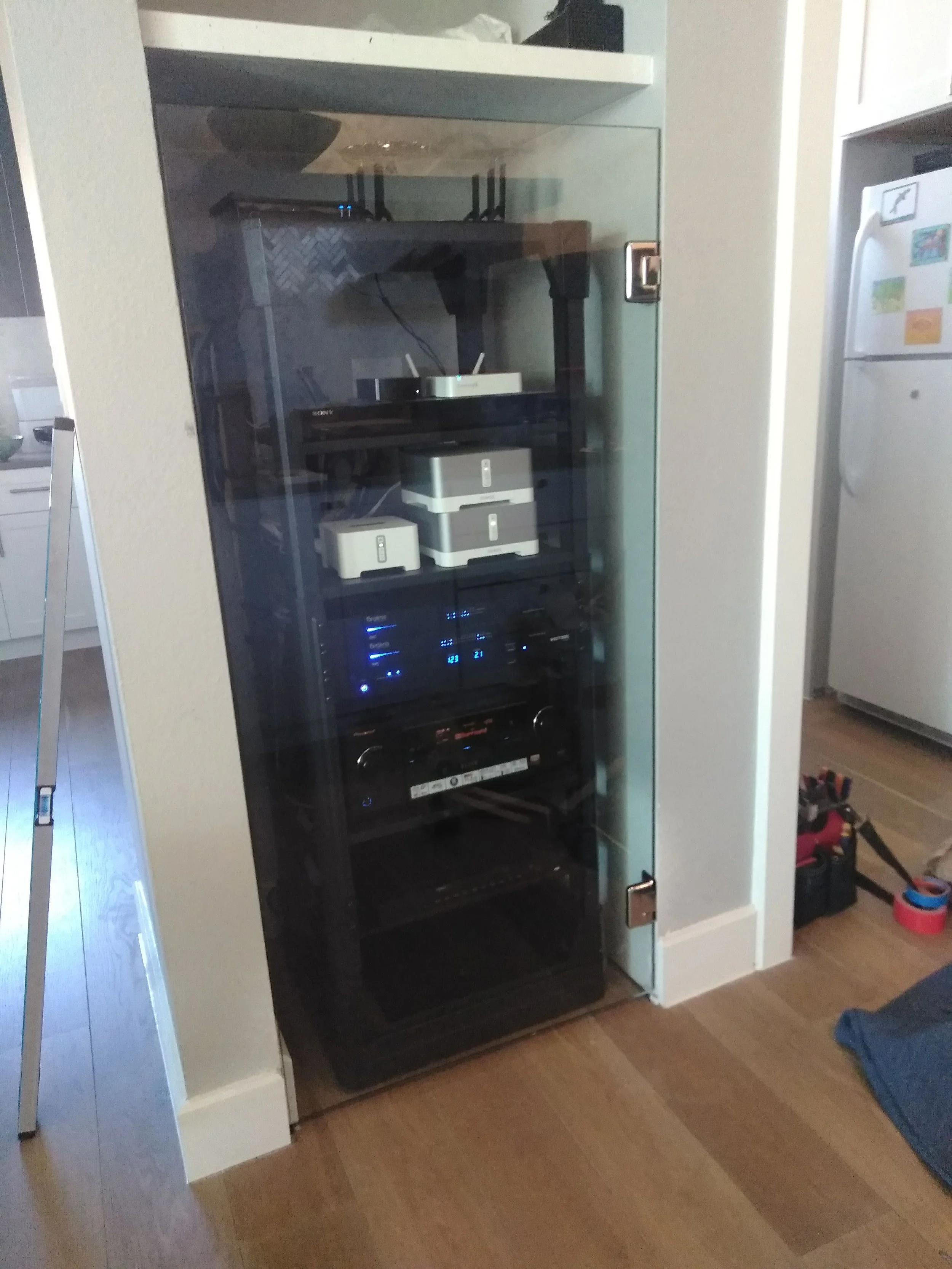 A black server rack with glass door holding electronic audio equipment and white containers, located against a white wall in a room with wooden flooring.
