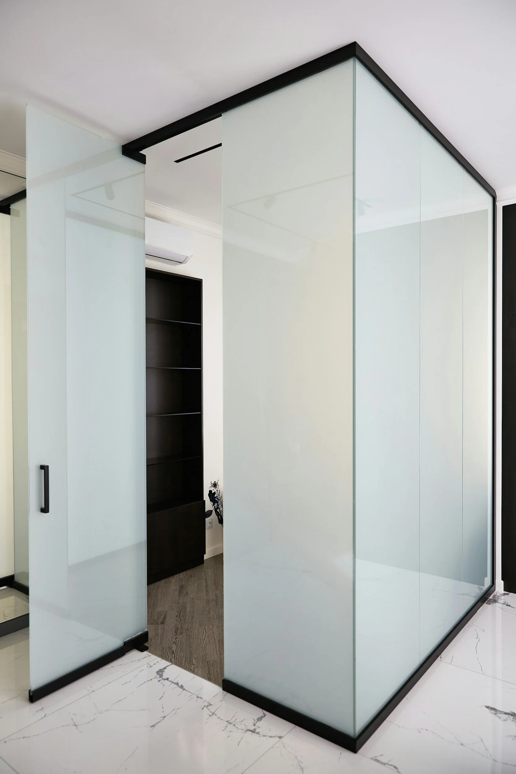 Modern glass partition wall with black frame surrounding a doorway and a walk-in closet in the background, situated in a room with white marble flooring.