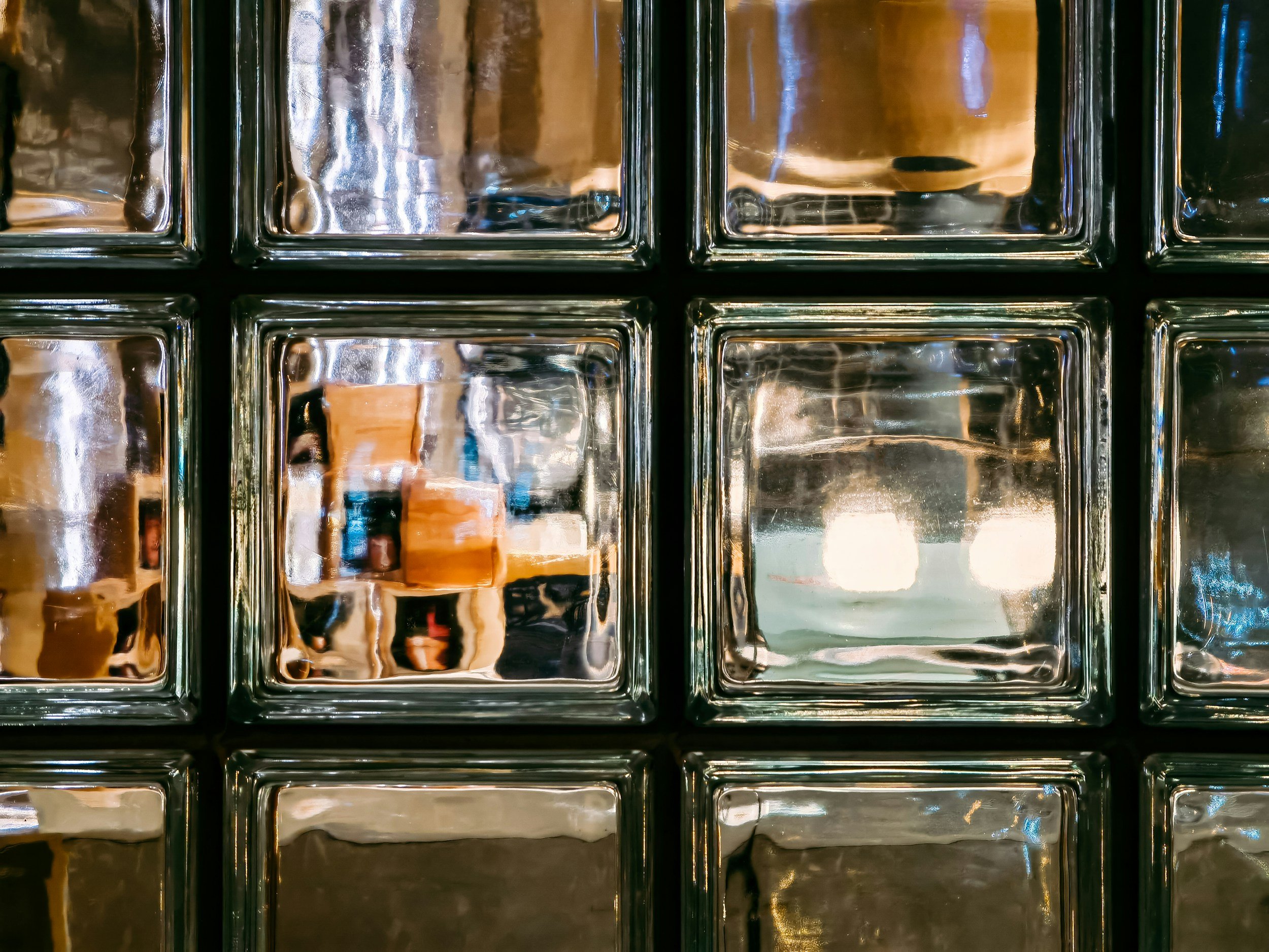 A close-up view of glass bricks or tiles with reflective surfaces and dark grout lines, showing distorted reflections of objects and surroundings.