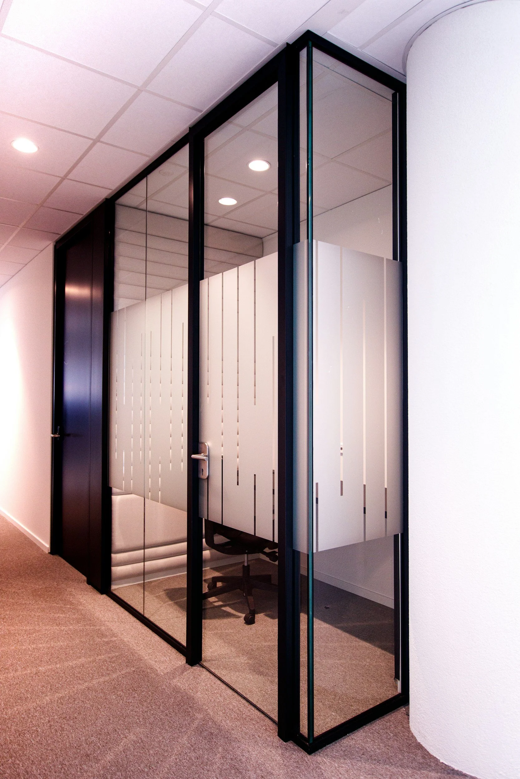 Modern glass-walled office partition with a white desk inside and a black office chair. The partition has a black metal frame and frosted glass panels with vertical lines, dividing the space from the hallway with beige carpet and white walls.