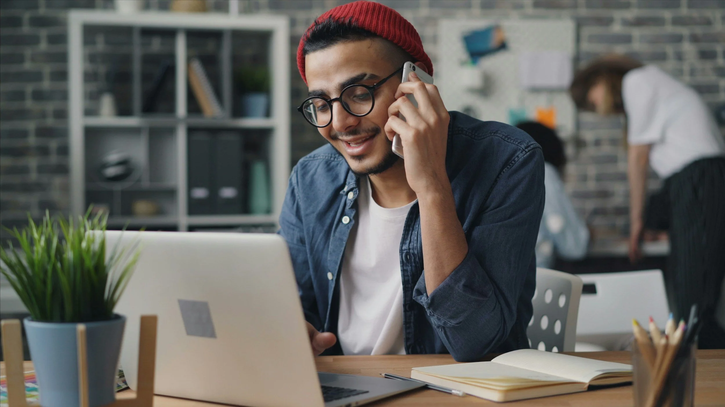 A young man with glasses and a red beanie talking on the phone while working at a desk with a laptop, notebook, and a potted plant in a modern office setting.