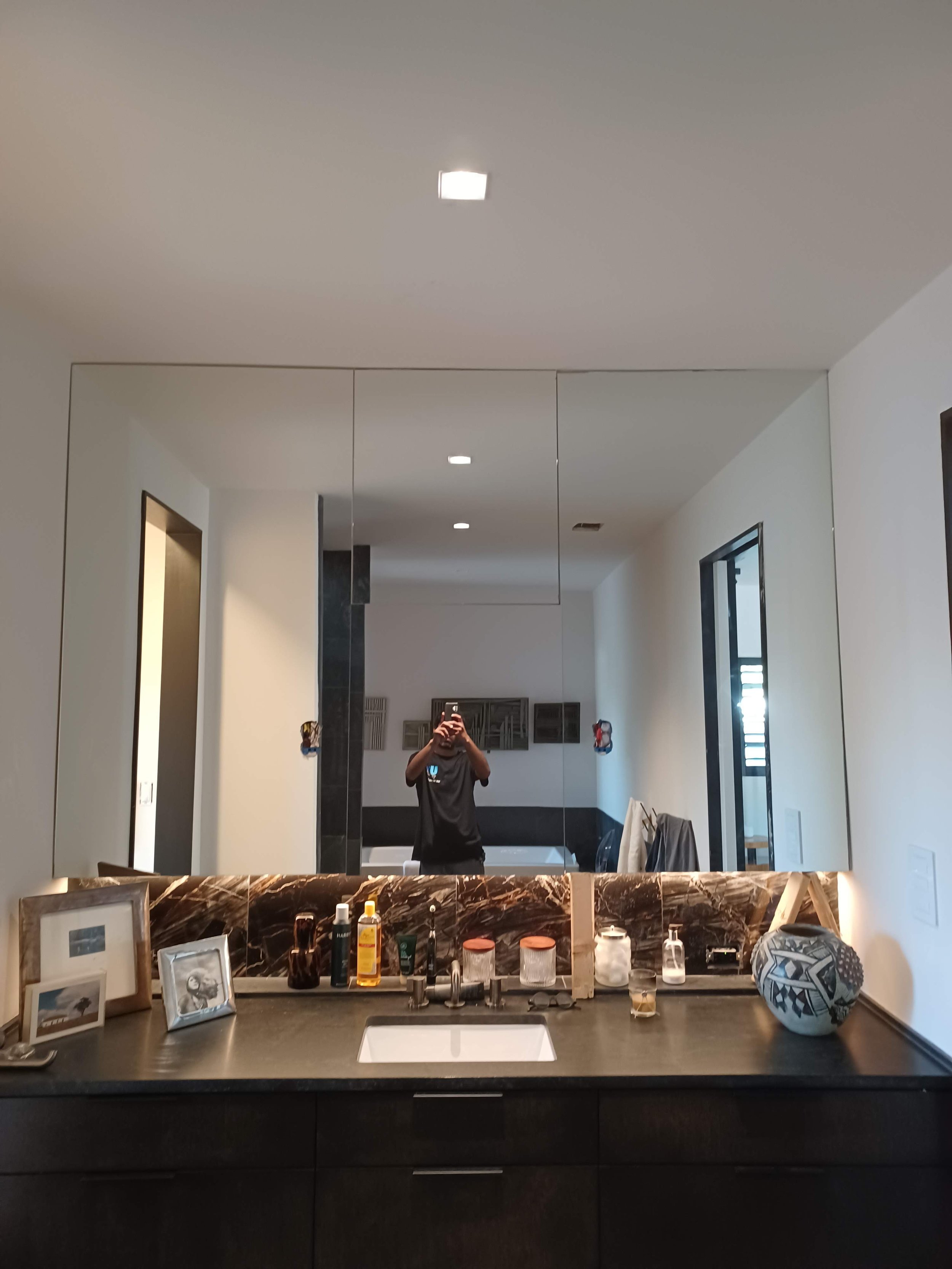 A bathroom vanity mirror reflects a person taking a photo with a phone, surrounded by bathroom items including framed photos, toiletries, and decorative objects on a marble countertop.