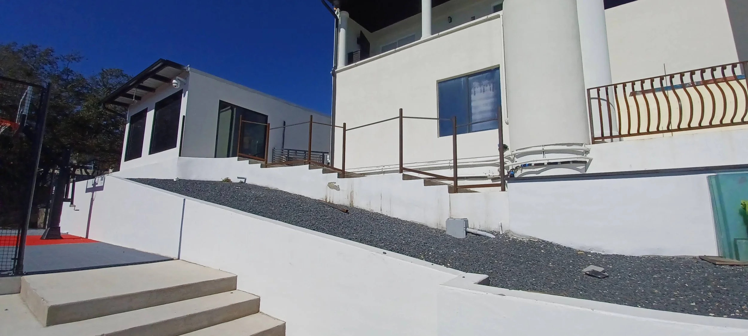 Modern white multi-story house with balconies, glass doors, and metal railings, situated on a hill with gray gravel and concrete steps leading to an outdoor area.