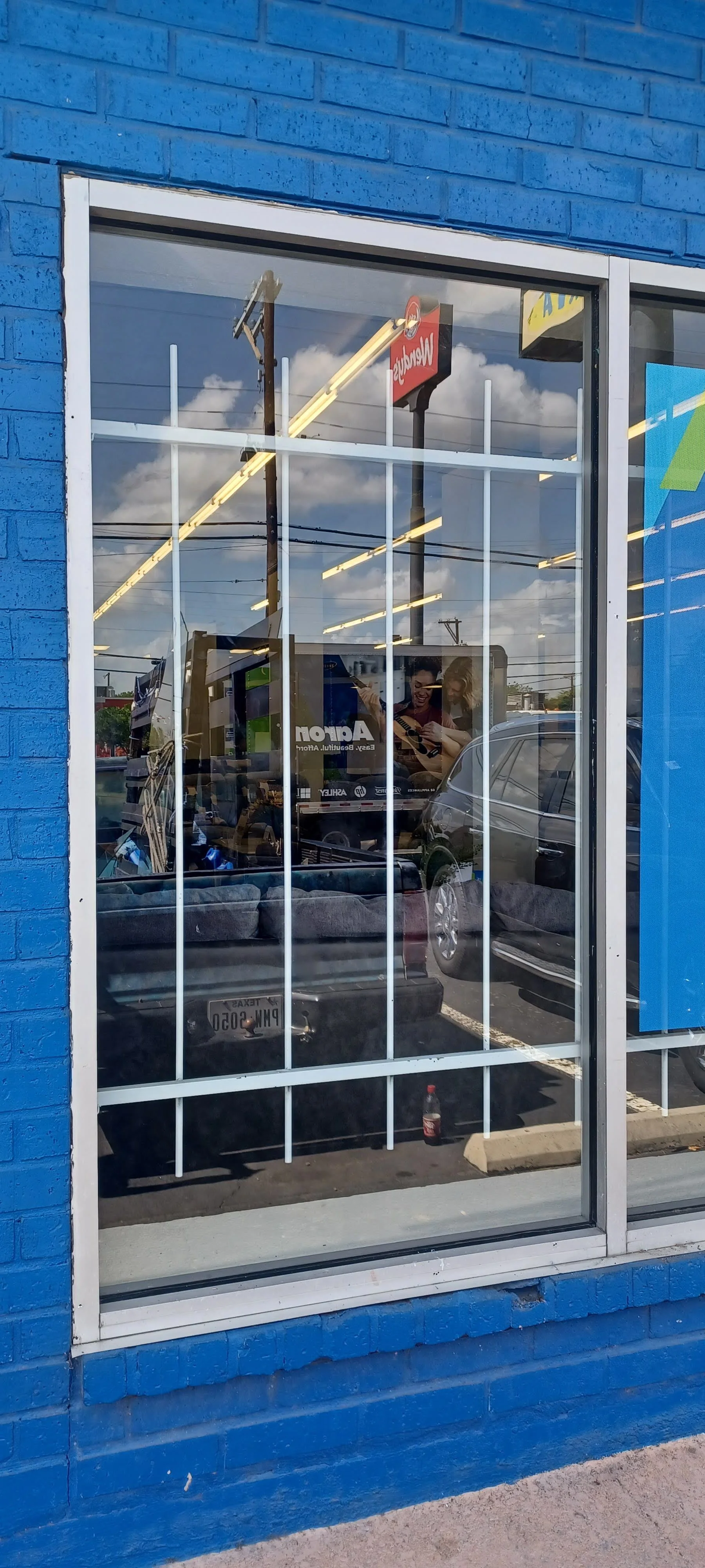 Reflected view of a parking lot through a window of a blue-painted building, showing parked cars, a billboard, and a cloudy sky.