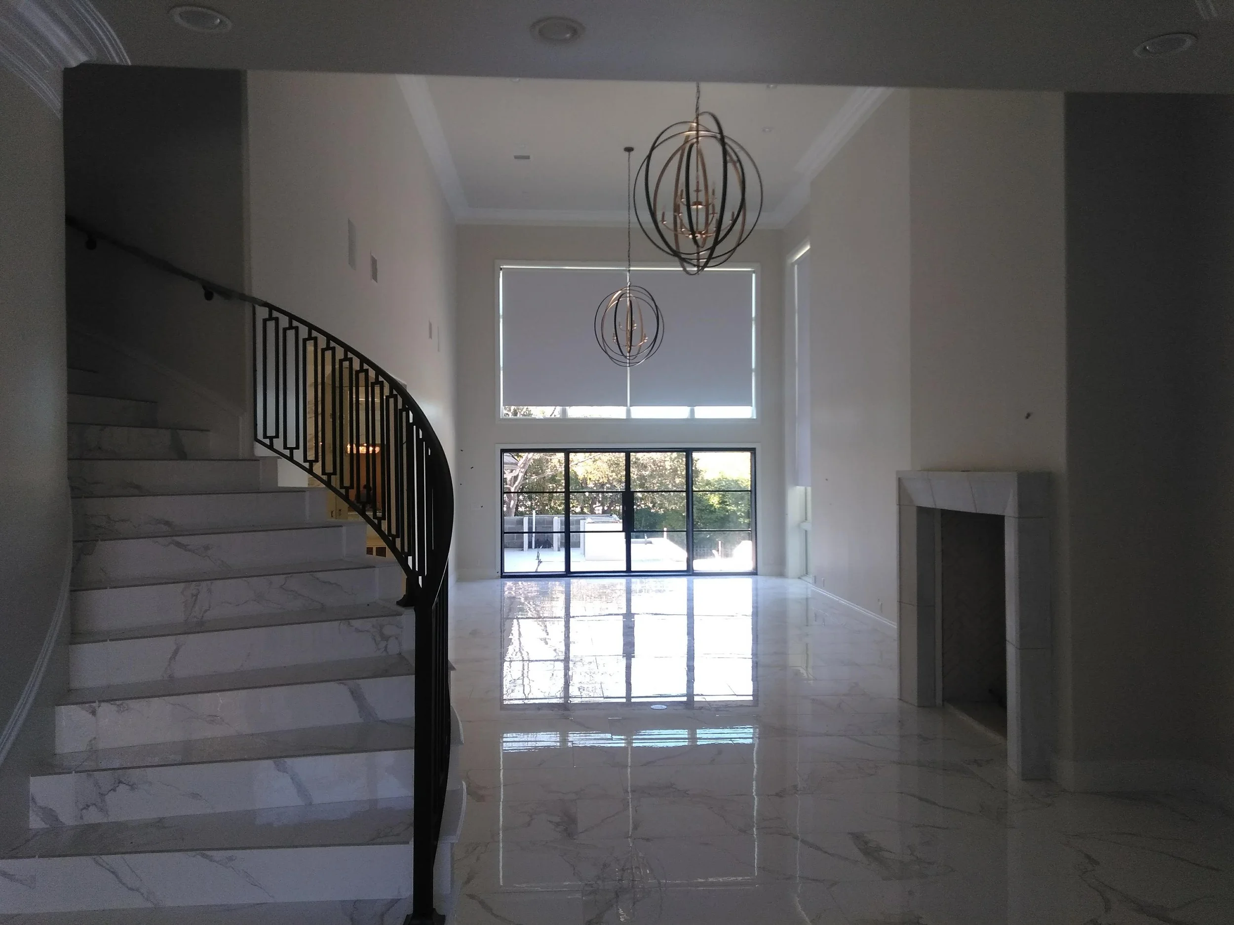 Interior view of a modern house with marble flooring, a staircase with metal railing, and large windows letting in natural light, showcasing a spacious and bright living area.