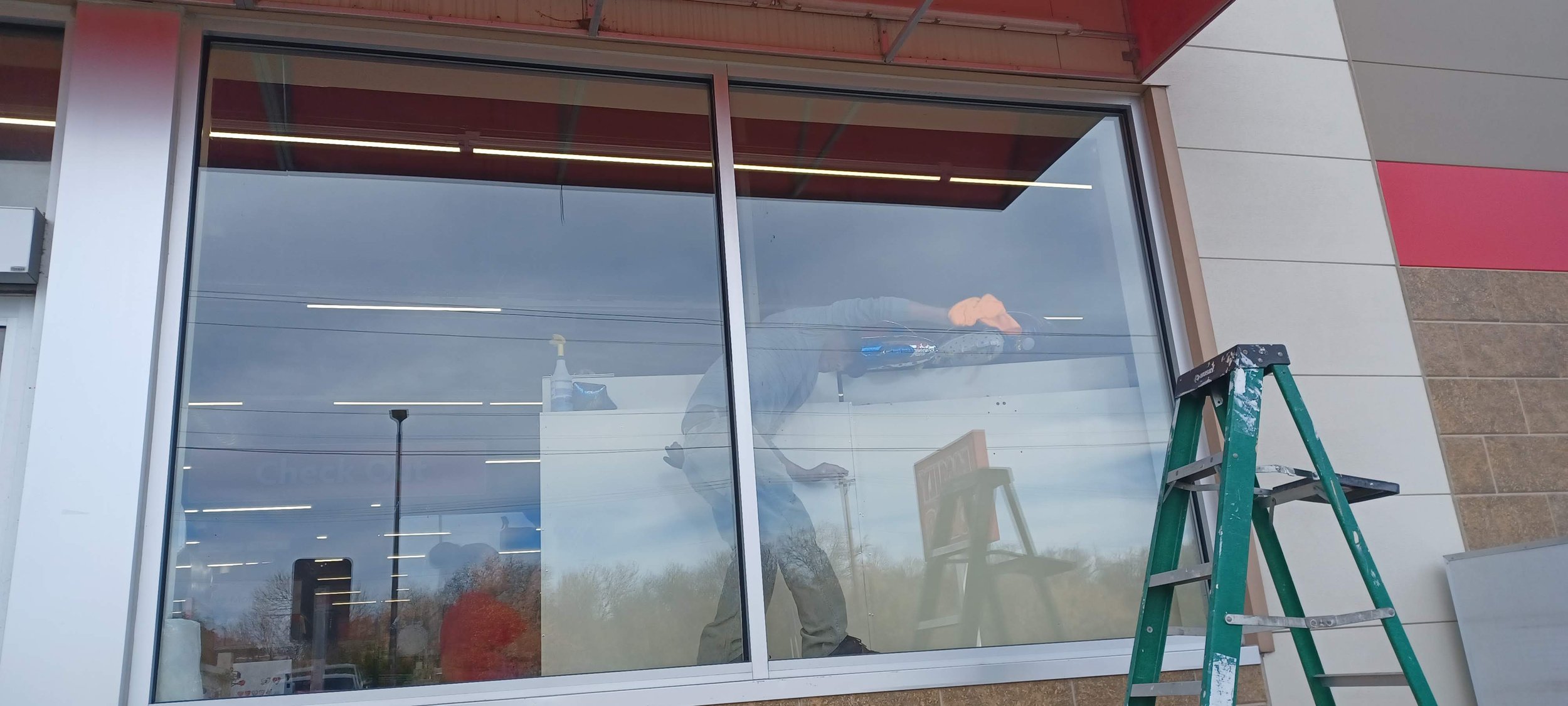 Person installing or maintaining a large glass window or storefront, standing on a ladder inside the building, with tools and cleaning supplies nearby.