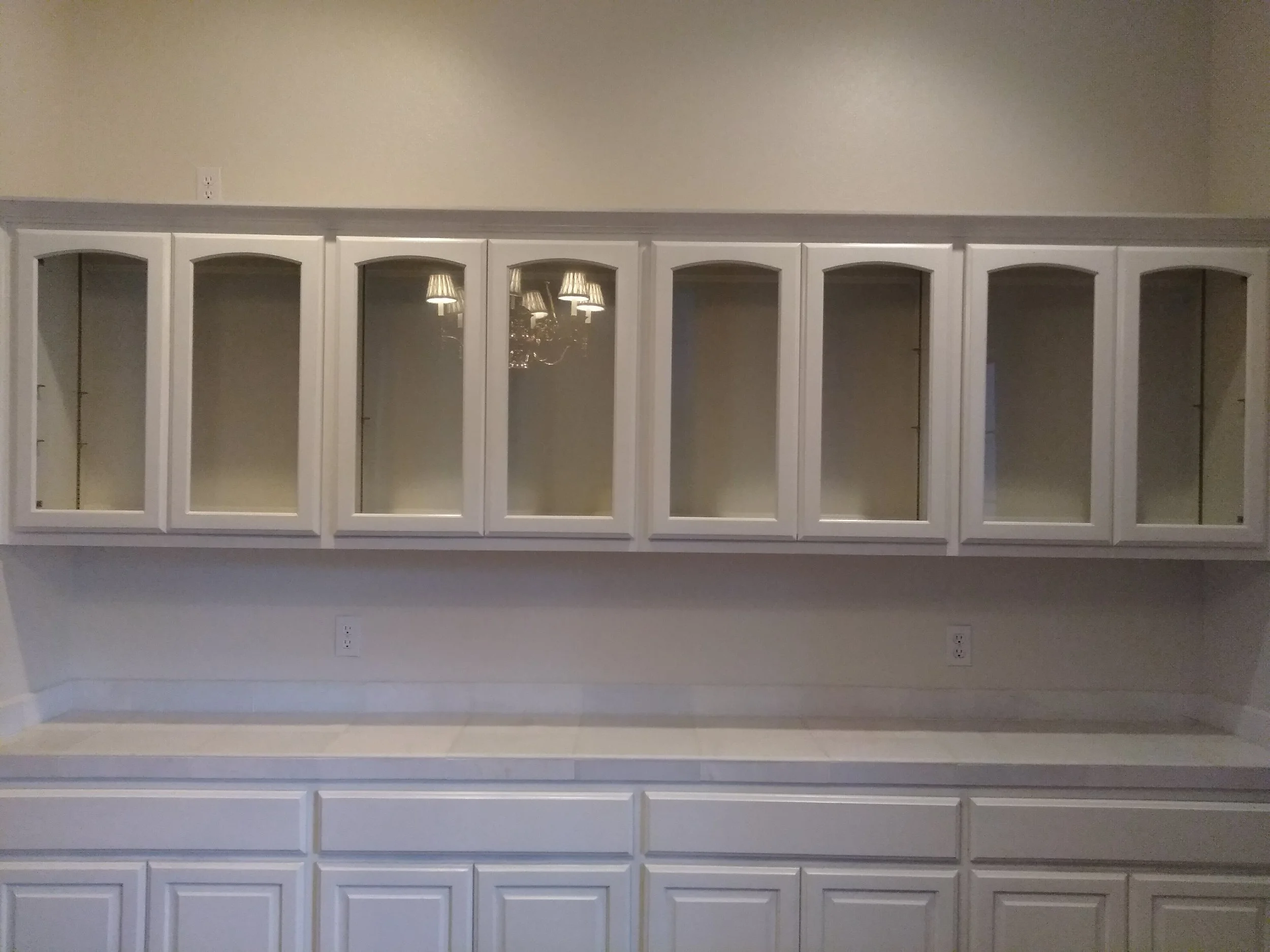 A built-in white cabinet with glass-paneled doors, interior shelves, and a reflection of chandeliers visible through the glass.