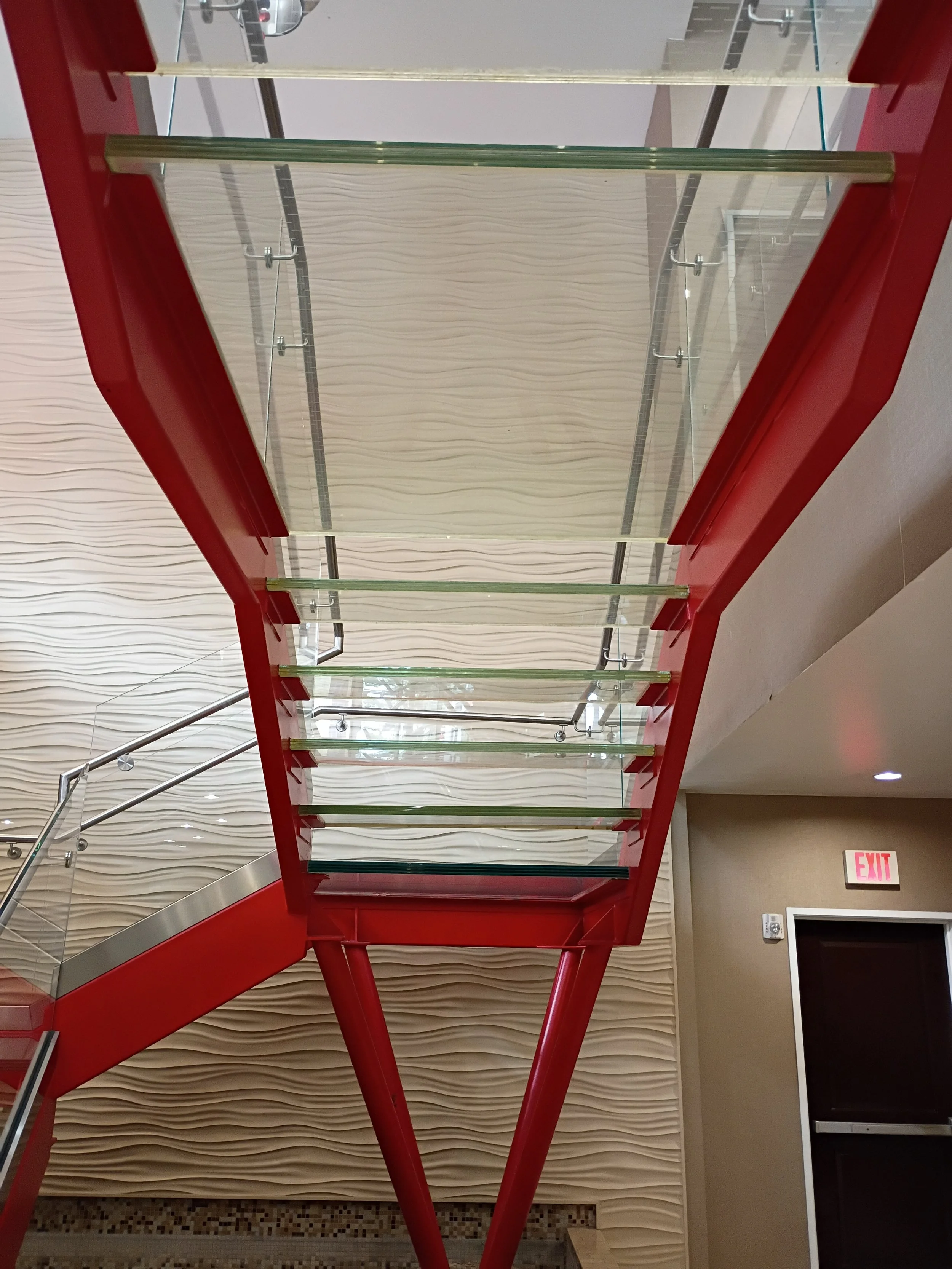 Red staircase with glass steps in an indoor setting, with a textured beige wall behind and an exit sign on the door to the right.
