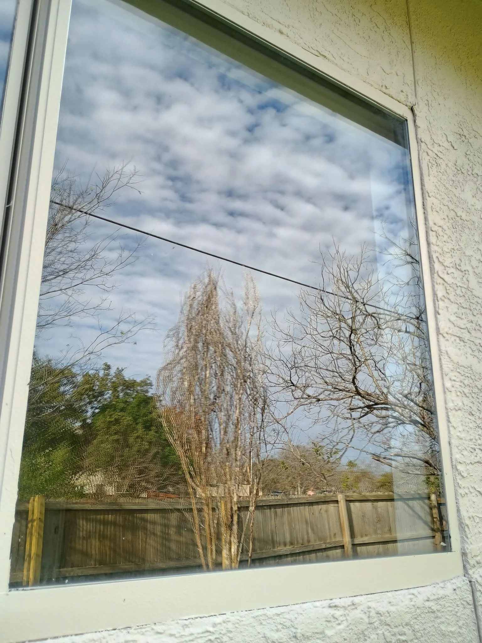 View of a partially visible window reflecting a cloudy sky, leafless trees, and a wooden fence outside.