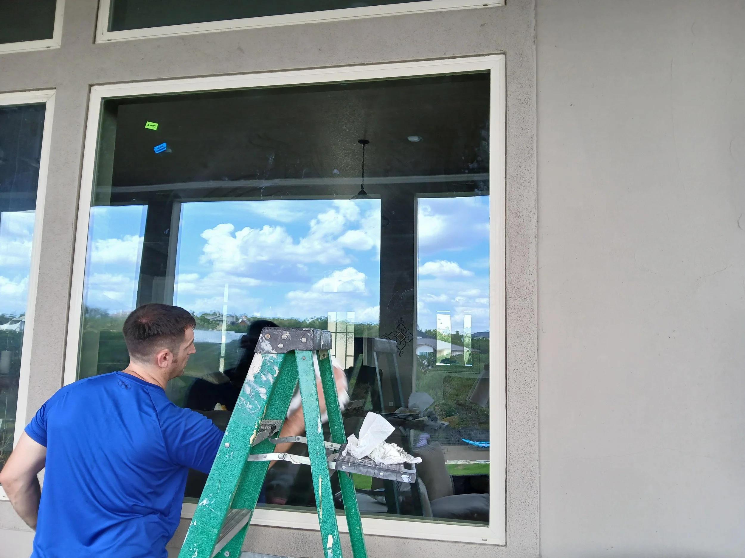 A man in a blue shirt working on repairing or installing a window from a ladder outside a house. The window reflects a cloudy sky and nearby landscape.