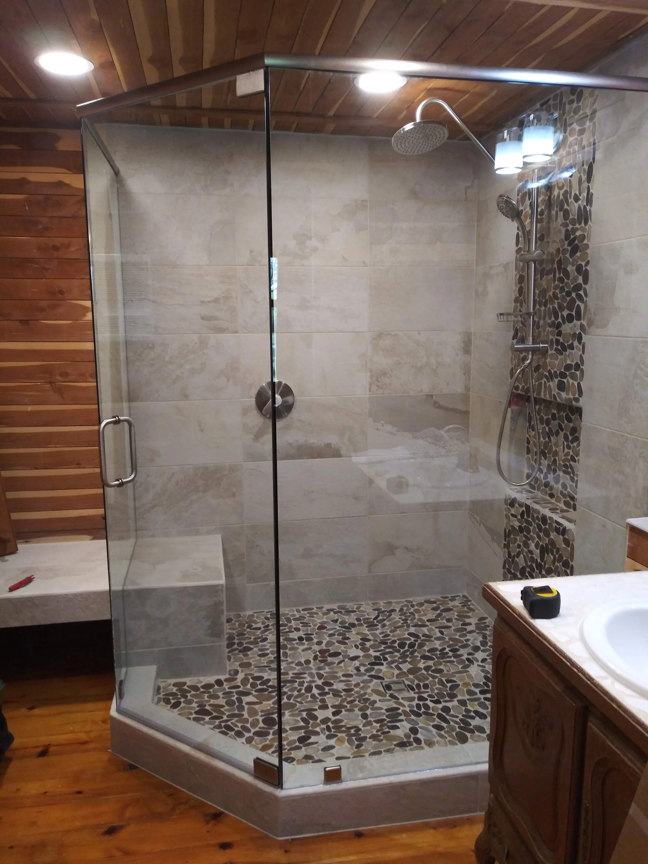A modern walk-in shower with a glass door, pebble tile floor, beige tiled walls, and a showerhead. There is a wood-paneled ceiling and a wooden cabinet is partly visible to the right.