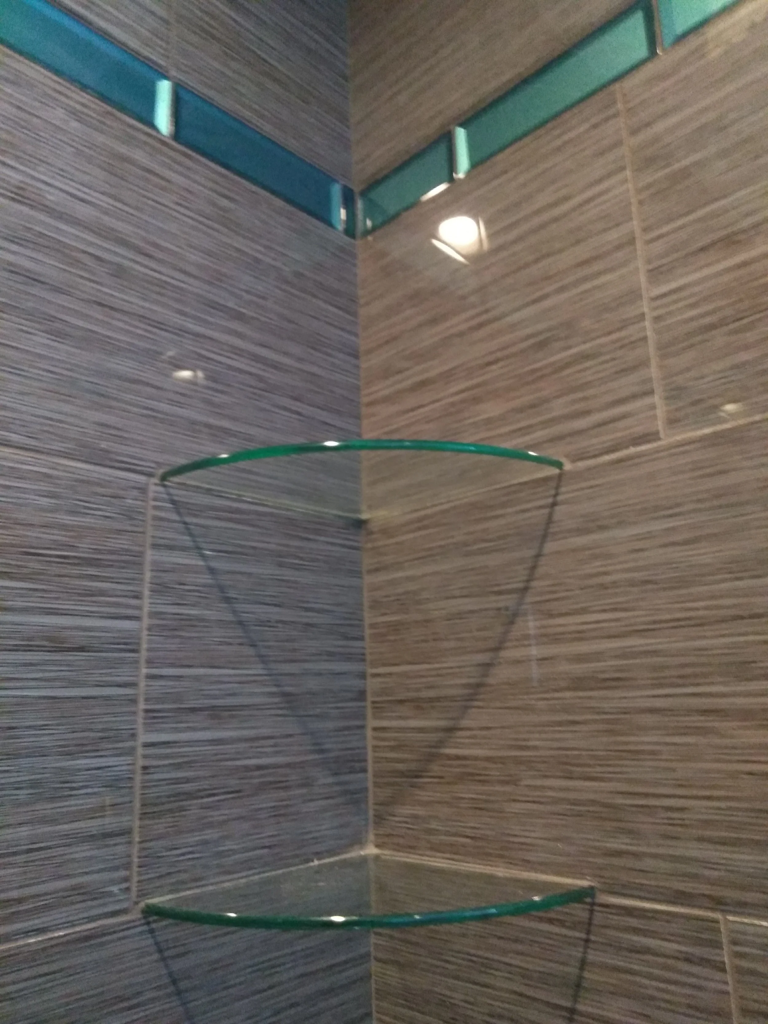 Corner of a shower with gray and beige striped tiles, a small glass shelf, and water reflection on the tiles.