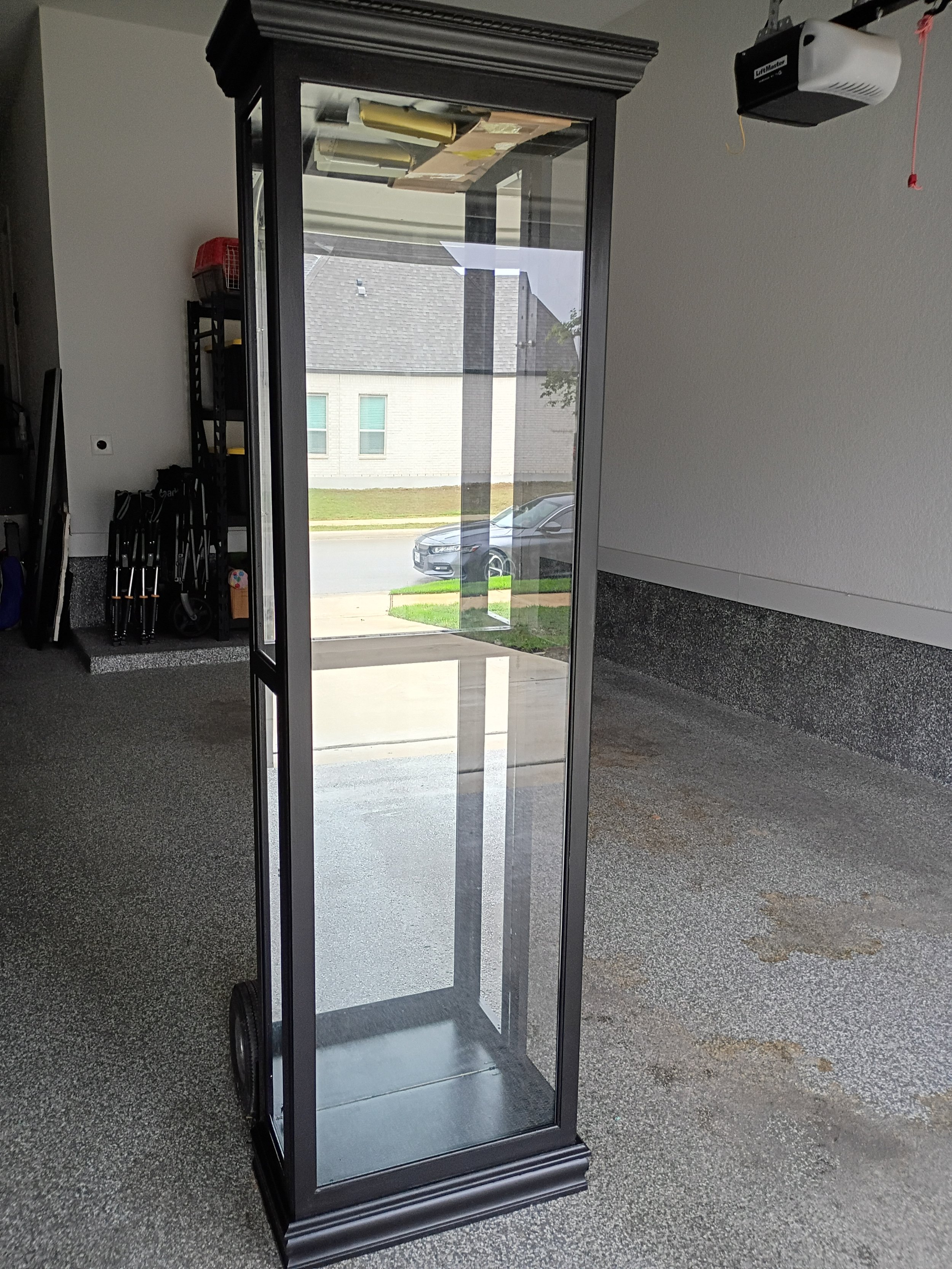 A tall, black, glass display cabinet with a decorative base, positioned on a textured gray carpet in a garage or storage space, with a partially open glass door showing a view of a street and parked cars outside.