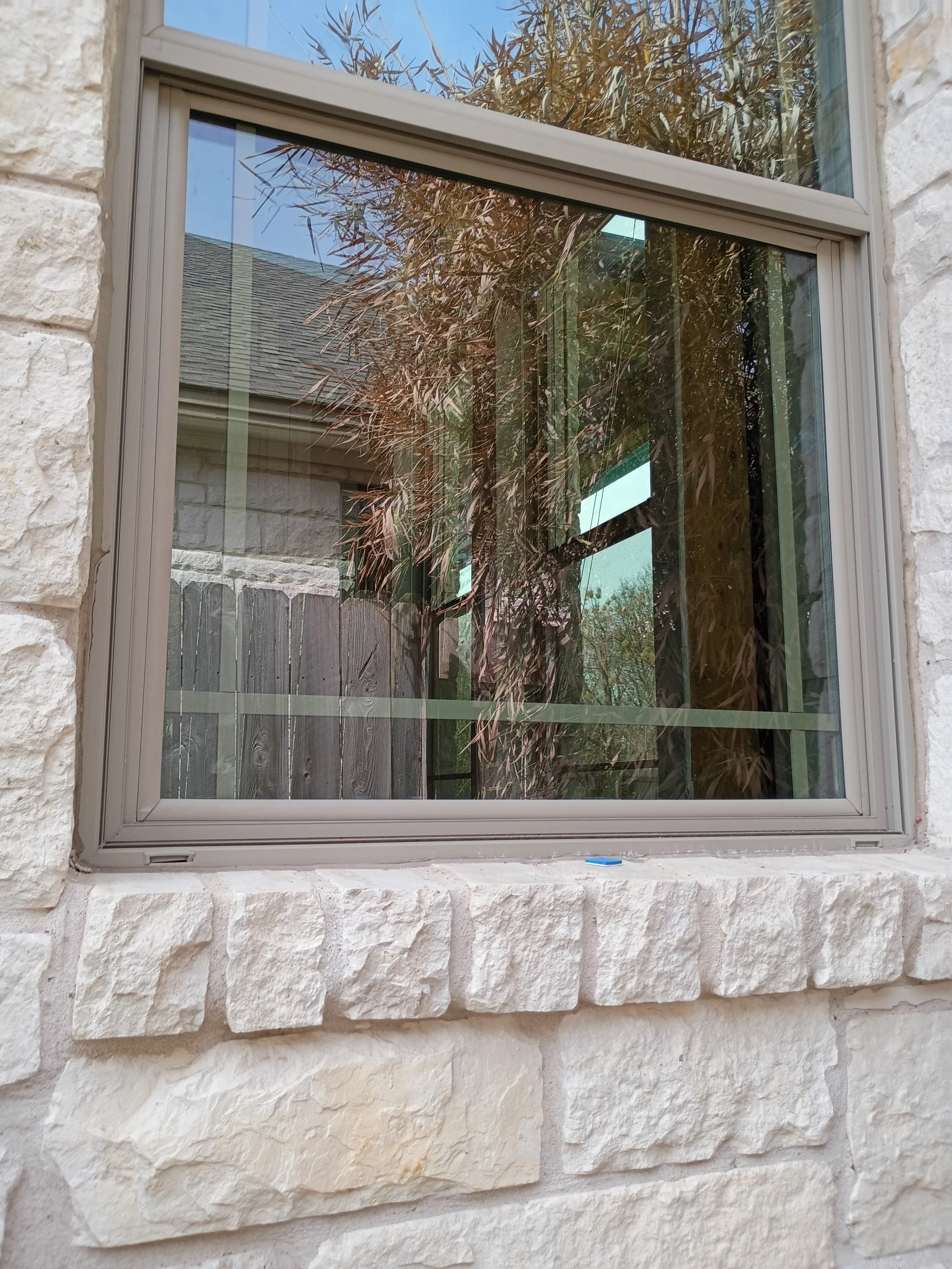 A window set in a stone wall. The window has a beige frame and reflects trees and part of a roof outside.