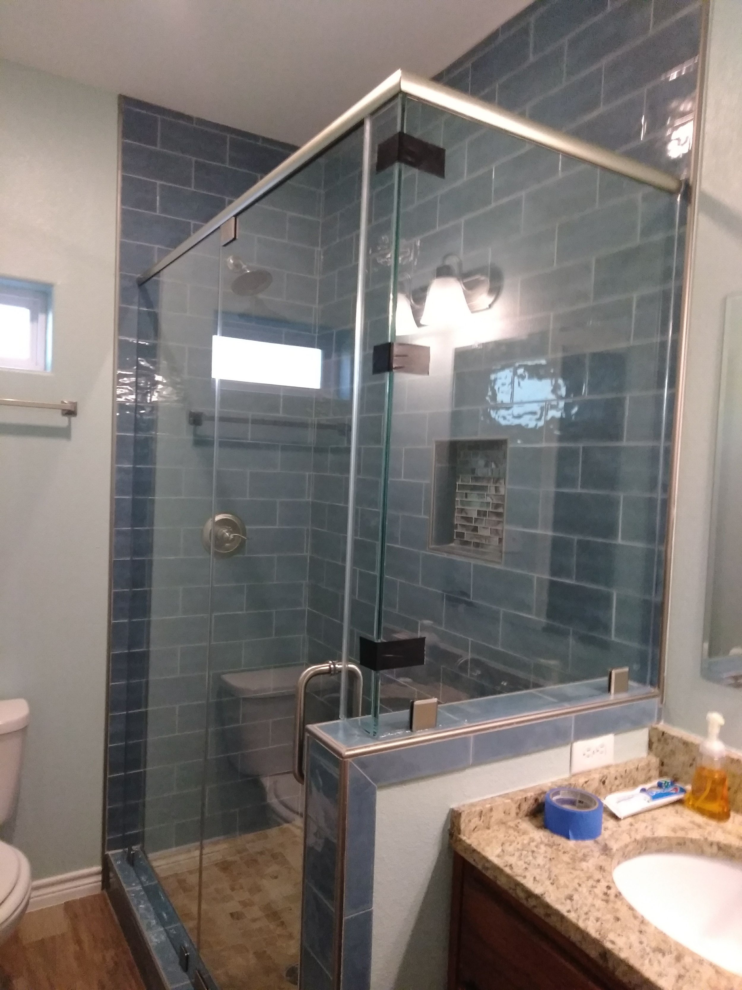 Bathroom with a glass shower enclosure featuring blue tiles, a meditative showerhead, a wood vanity with a granite countertop, a sink, and various toiletries and tools on the counter.