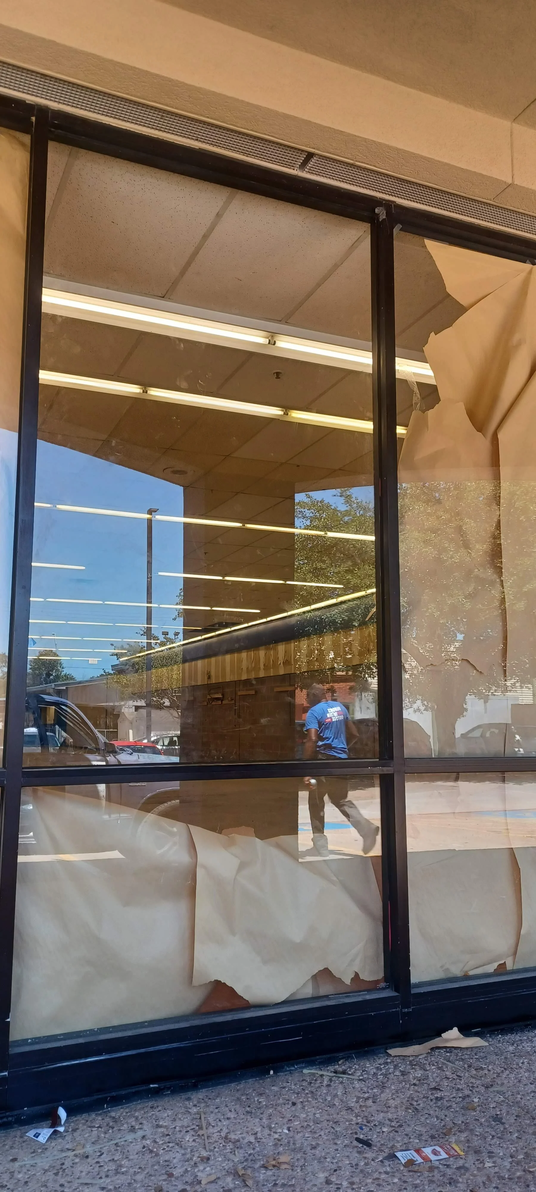 The photograph shows the exterior of a storefront with large glass windows. Inside, fluorescent lights are visible, and a person in a blue shirt is walking outside. The ground outside the shop has some trash and paper.