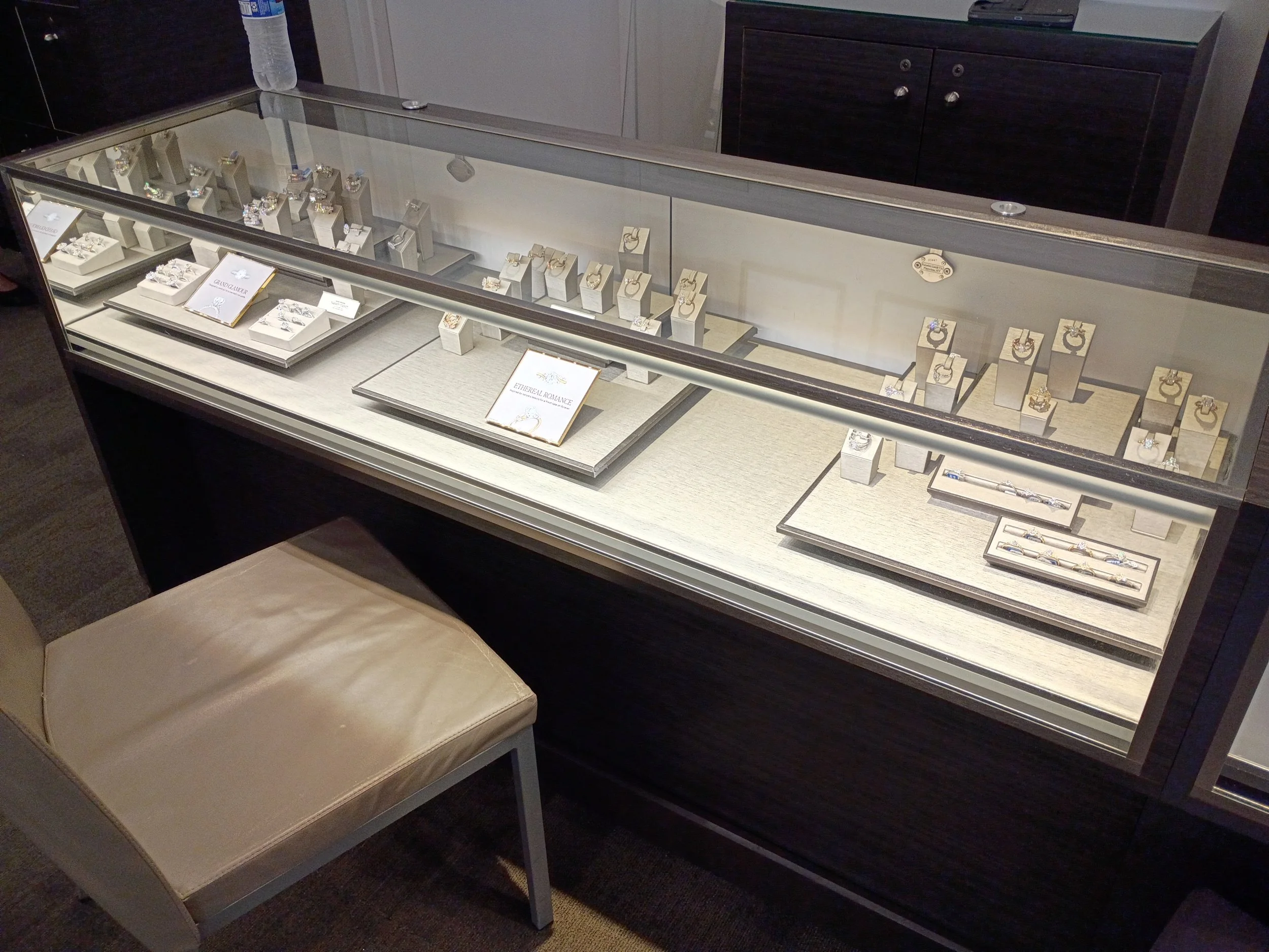 A jewelry display case showcasing rings and necklaces in a retail store. A beige cushioned chair is in front of the display.