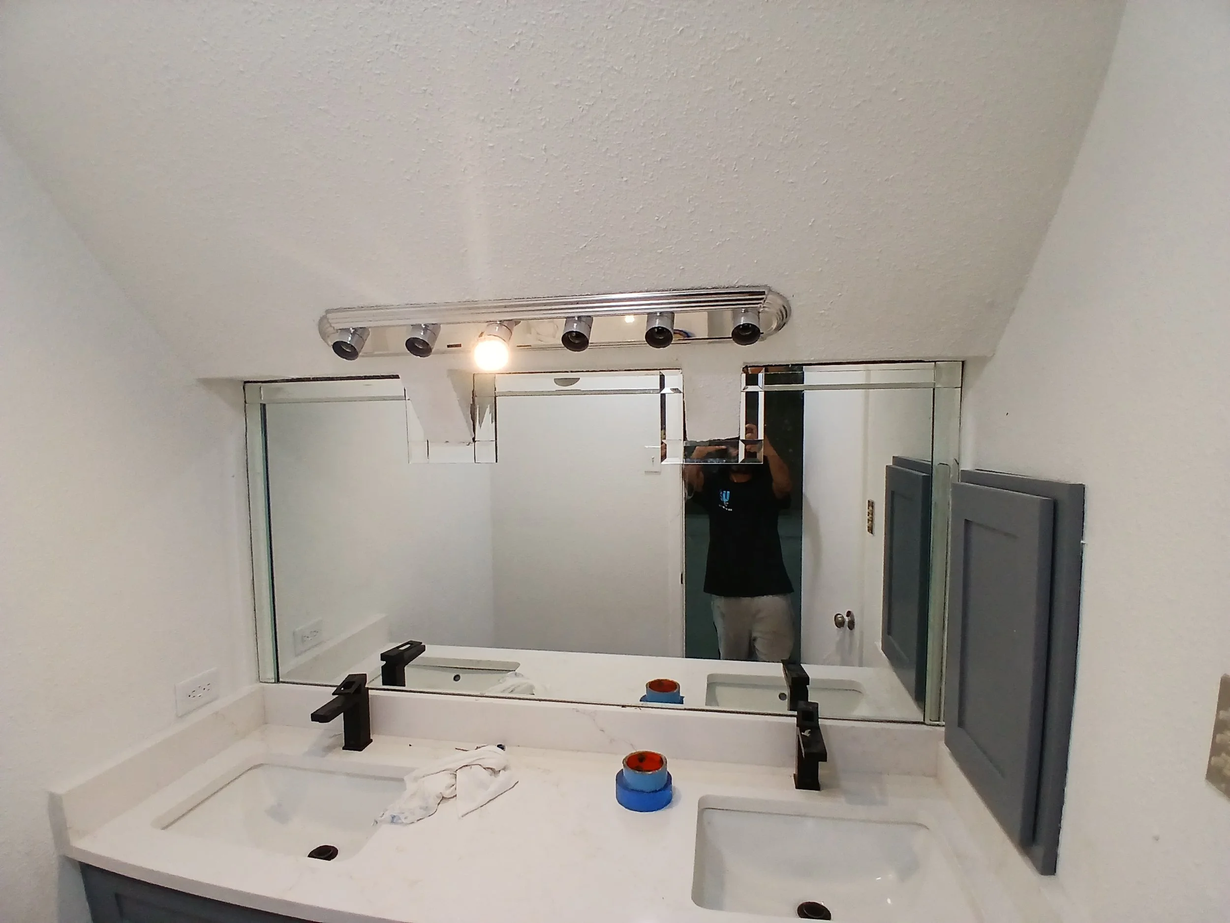 A bathroom under renovation with a large mirror, a white countertop with an under-mount sink, black fixtures, and unfinished cabinet or vanity. The wall above the countertop is being prepared for mirror or backsplash installation, with visible electr