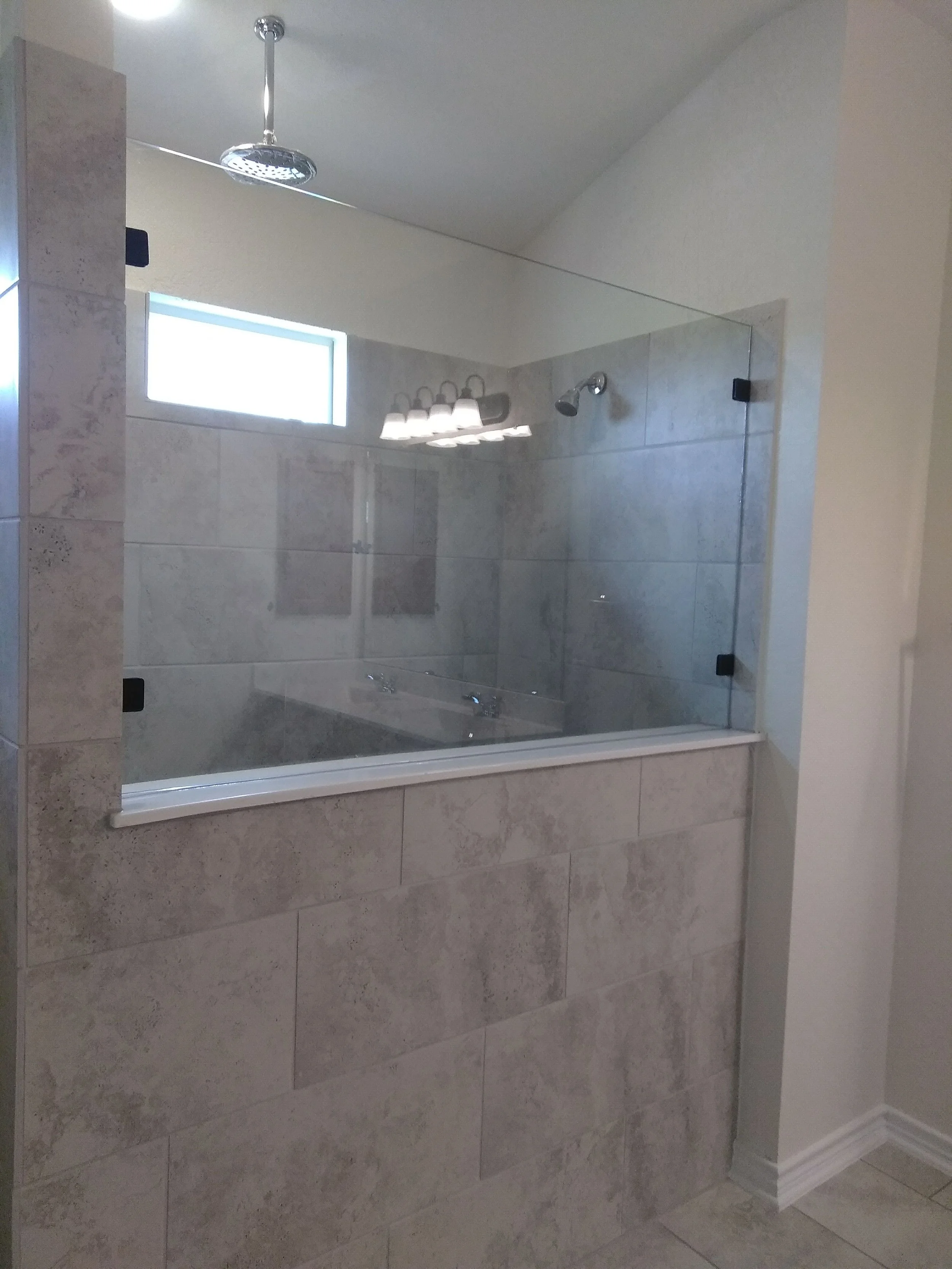 A bathroom with large beige tiled walls and a glass shower enclosure. Inside the shower, there's a window, a chrome showerhead, and a chrome shower for different hands-free and handheld use. The bathroom appears to be well-lit.