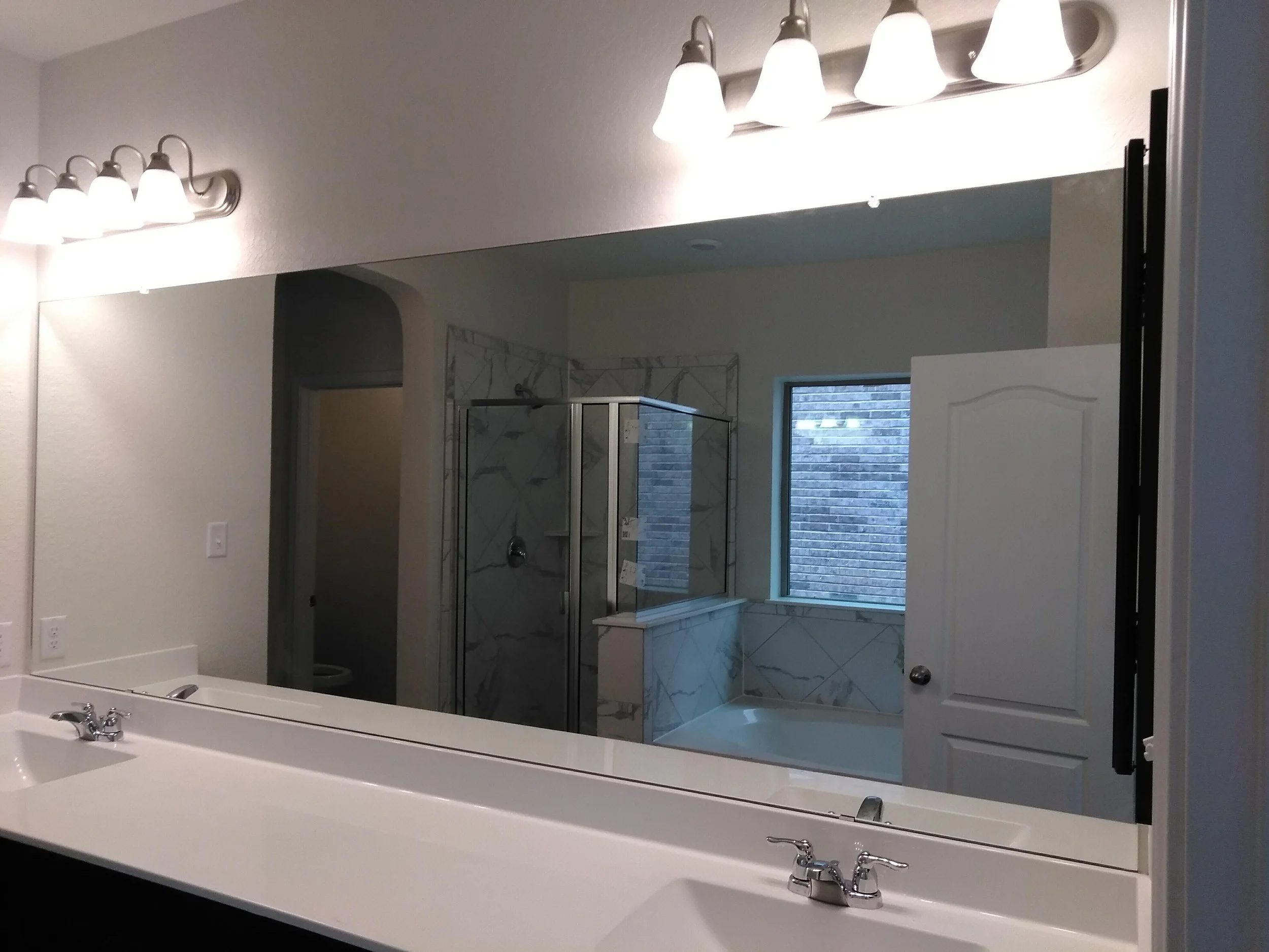 A bathroom with a large mirror reflecting a shower with a glass door, a window, and a white countertop with sinks and faucets.