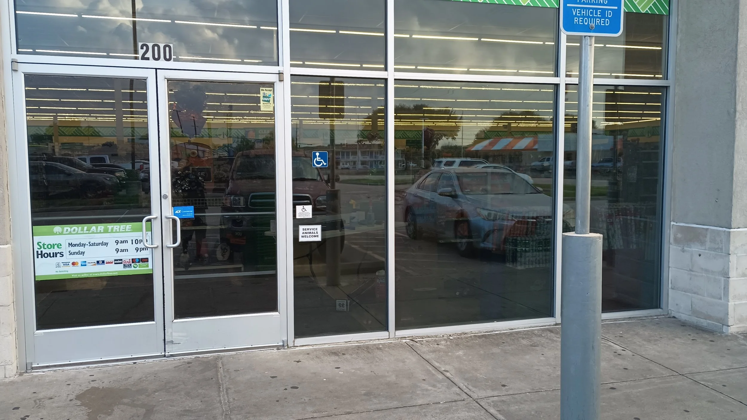Front glass entrance doors of a grocery store with reflections of cars and trees outside. Signs on the doors show store hours and acceptance of SNAP benefits, and a sticker indicates service animals are welcome. A handicapped parking sign is visible 