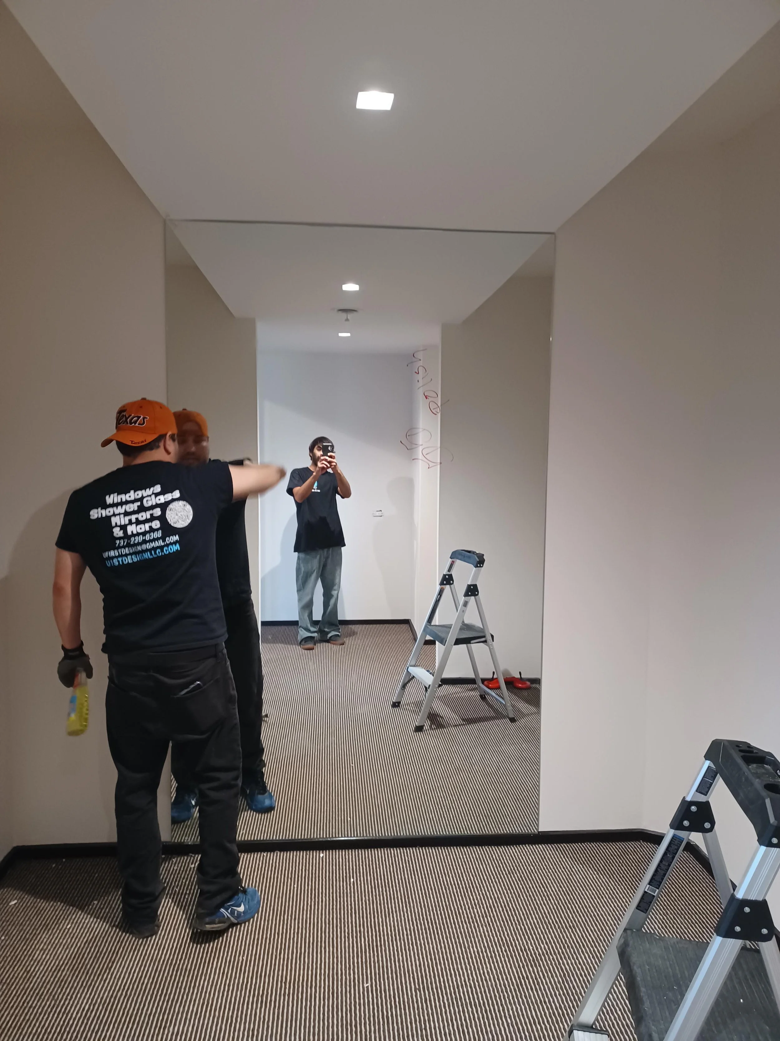 Three men working inside a hallway, with one taking a photo of the others in front of a large mirror. There is a ladder, tools on the floor, and writing on the wall.