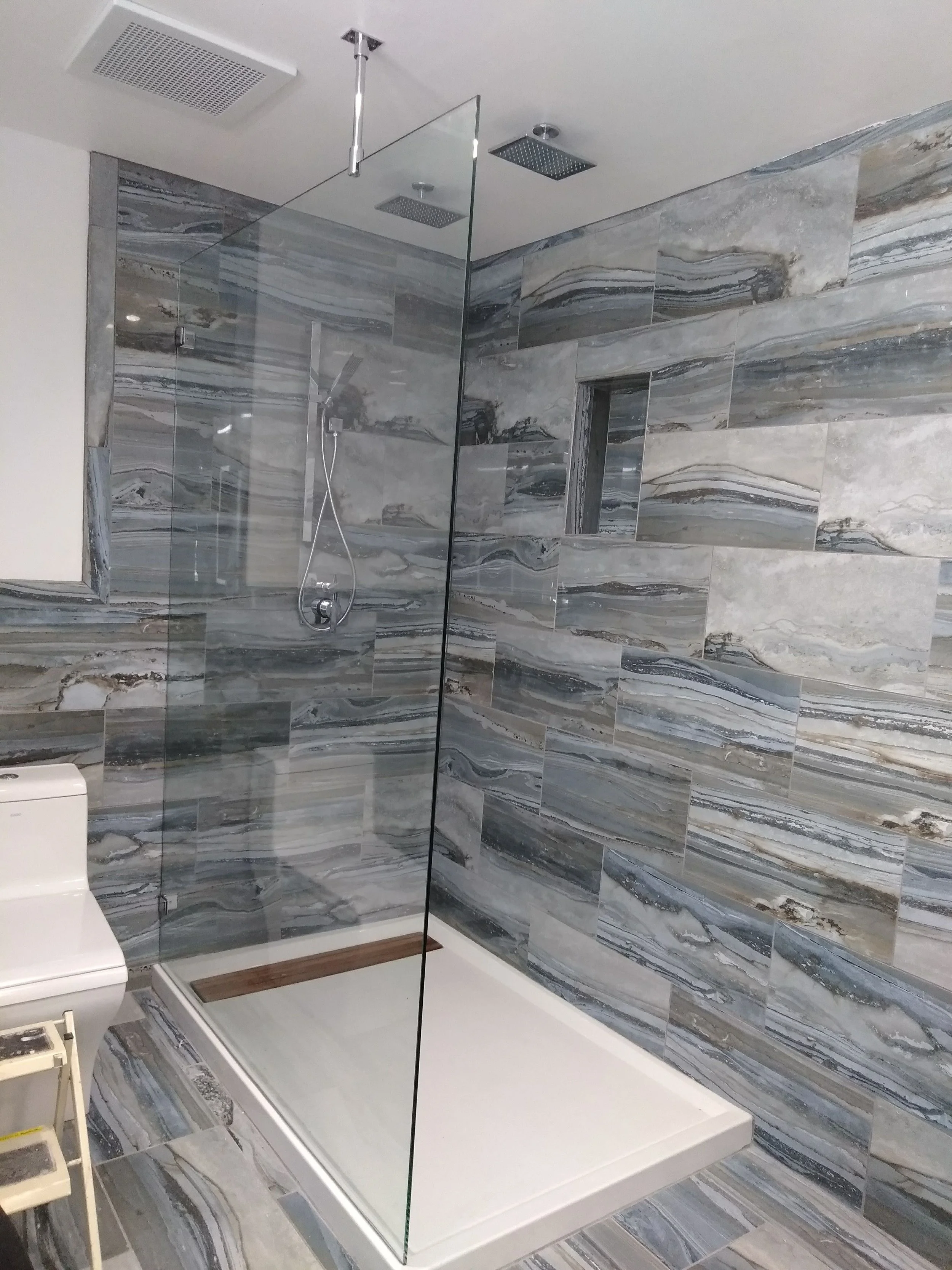 A modern walk-in shower with grey marble tiles, a glass partition, two showerheads on the ceiling, and a handheld shower on the wall. There is a built-in niche for toiletries and a white shower tray at the floor.