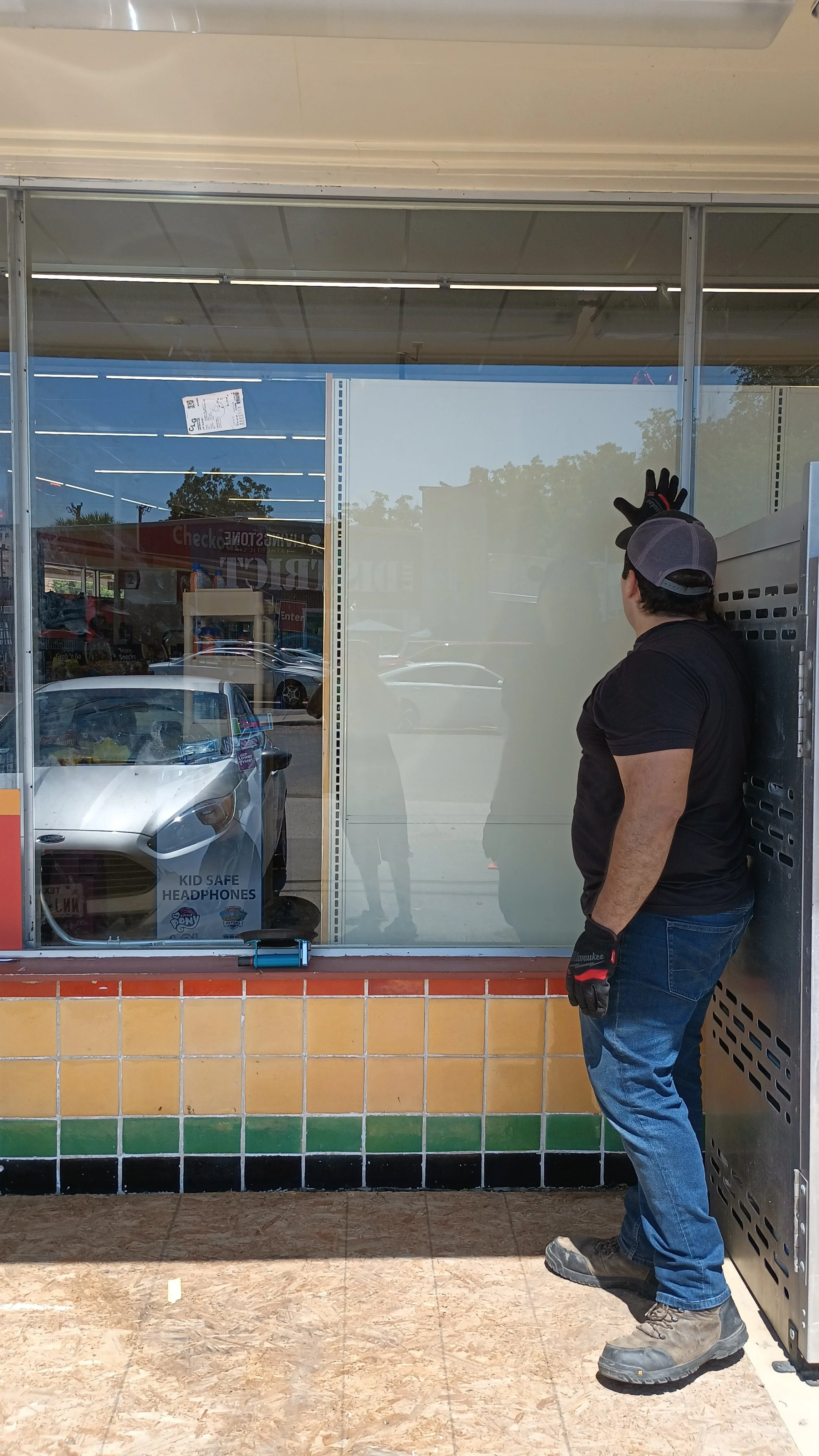 A man standing outside a store, wearing work gloves, a baseball cap, black t-shirt, jeans, and work boots, with his back turned, and looking through a large store window with reflections of cars and trees outside.