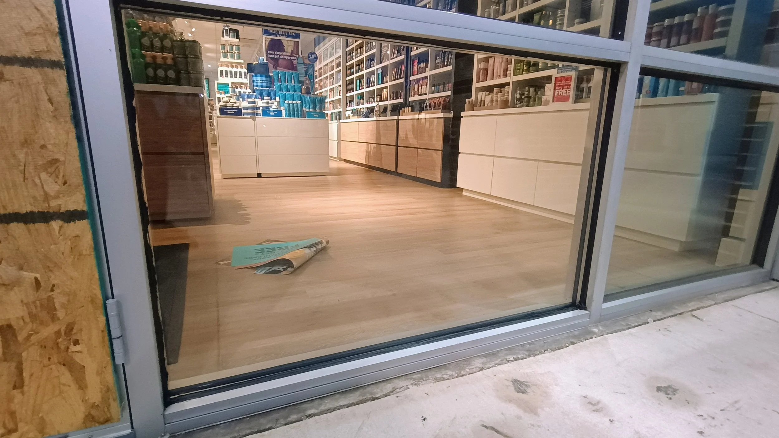 View of a store interior seen through a glass storefront. The store has wooden shelves and display cabinets with various products, including bottles and containers, and a wooden floor. There's a small wooden stand and a rolled newspaper on the floor 