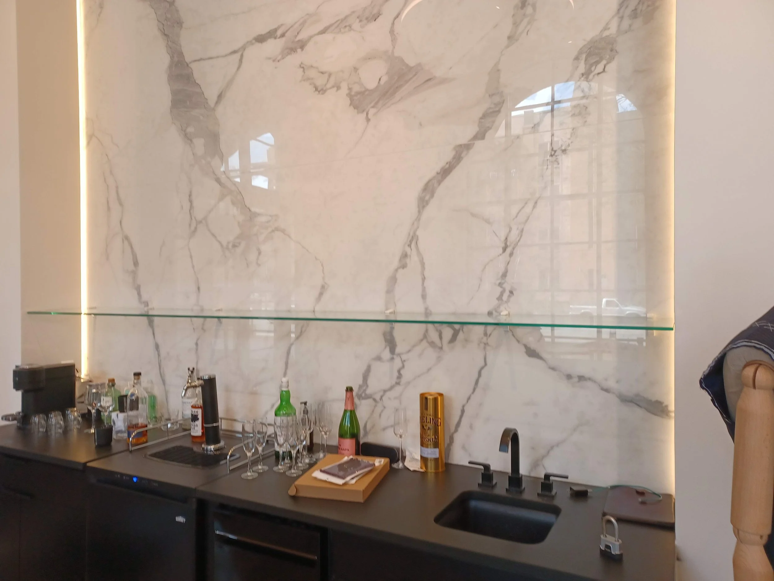 A modern bar with a marble backsplash behind it, various bottles, glasses, and bar tools on the counter, and some sunlight reflections on the marble surface.
