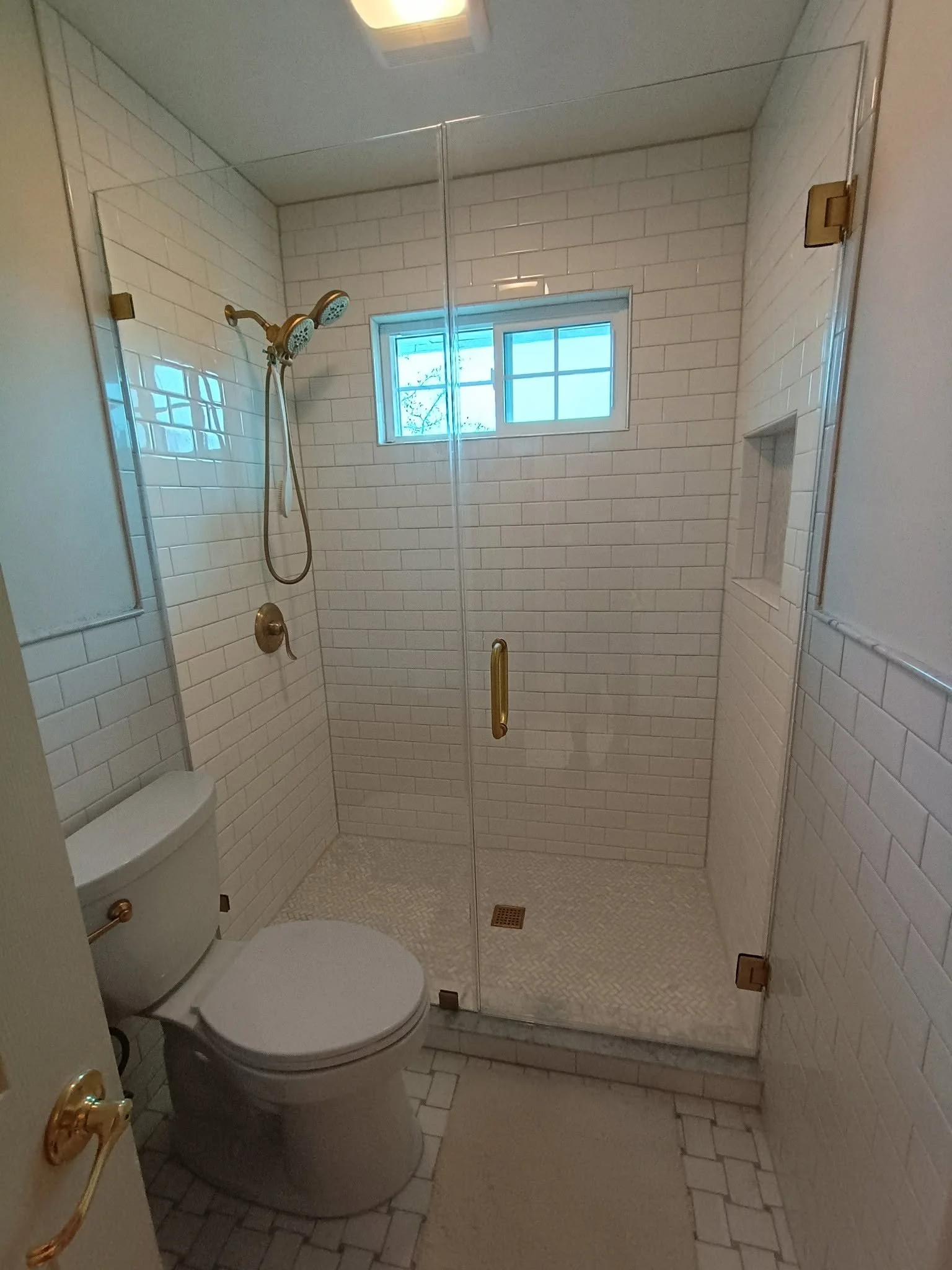 Small bathroom with white tiled walls, a toilet, a glass shower enclosure with a brass handle, dual shower heads, a small window, and a recessed shower shelf.