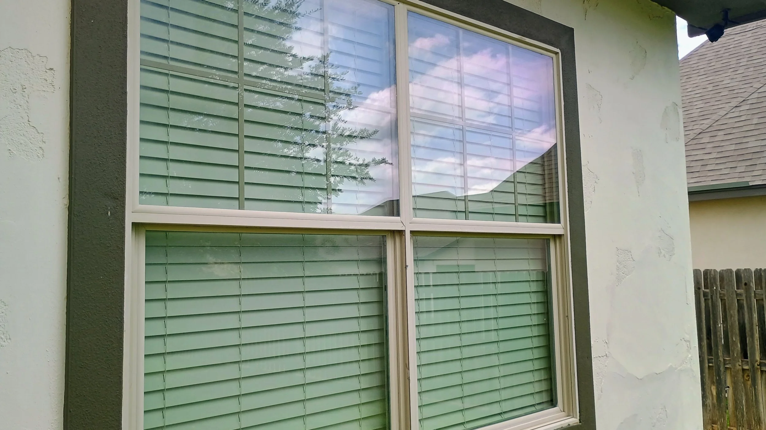 Close-up of a house window with closed green horizontal blinds, white window frame, and house wall with textured white paint and dark window trim. Reflection of trees and sky in the glass.