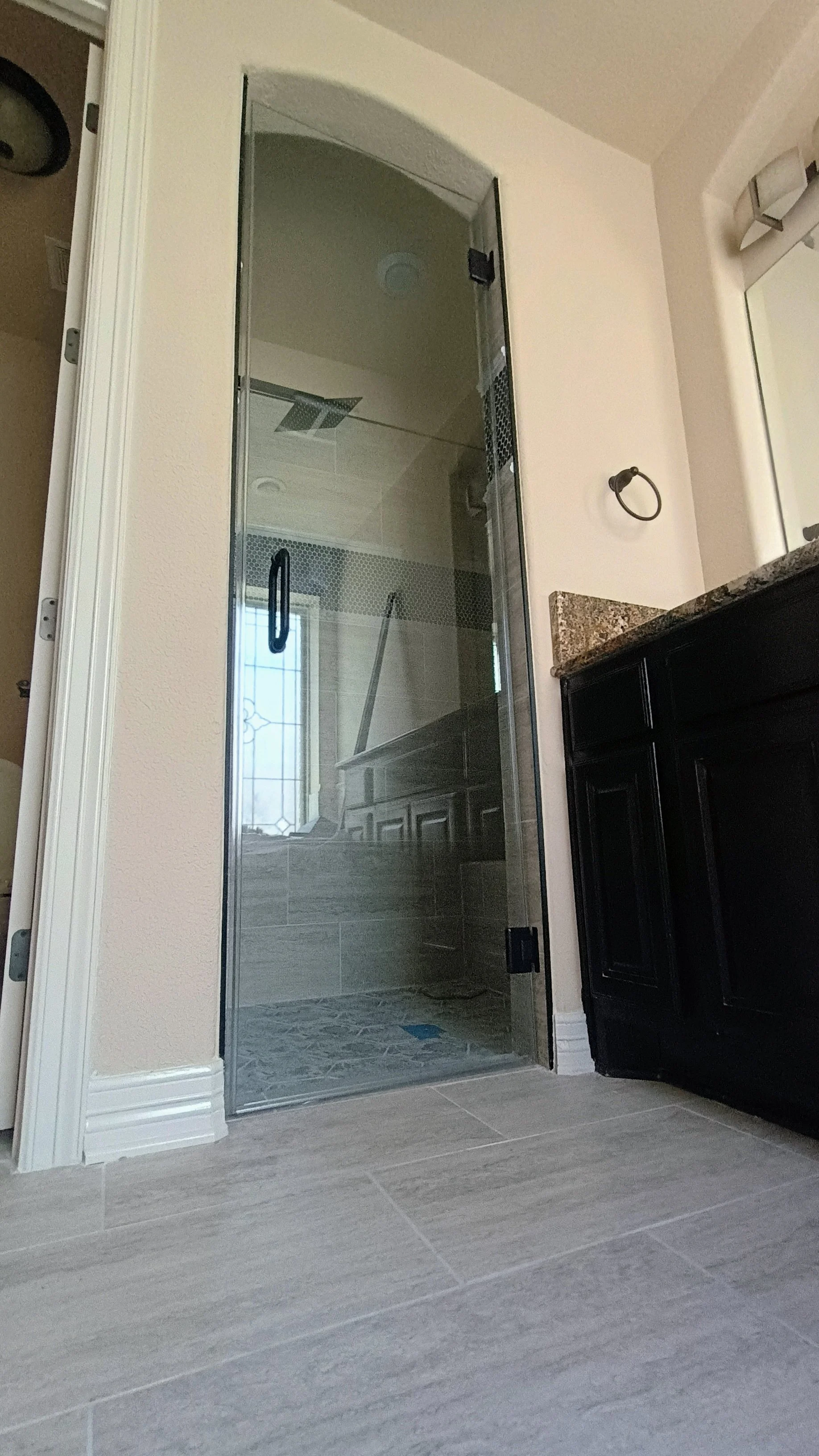 A bathroom with a glass shower door, beige tile floors, a dark wooden vanity with a granite countertop, and a wall-mounted light fixture above a mirror.