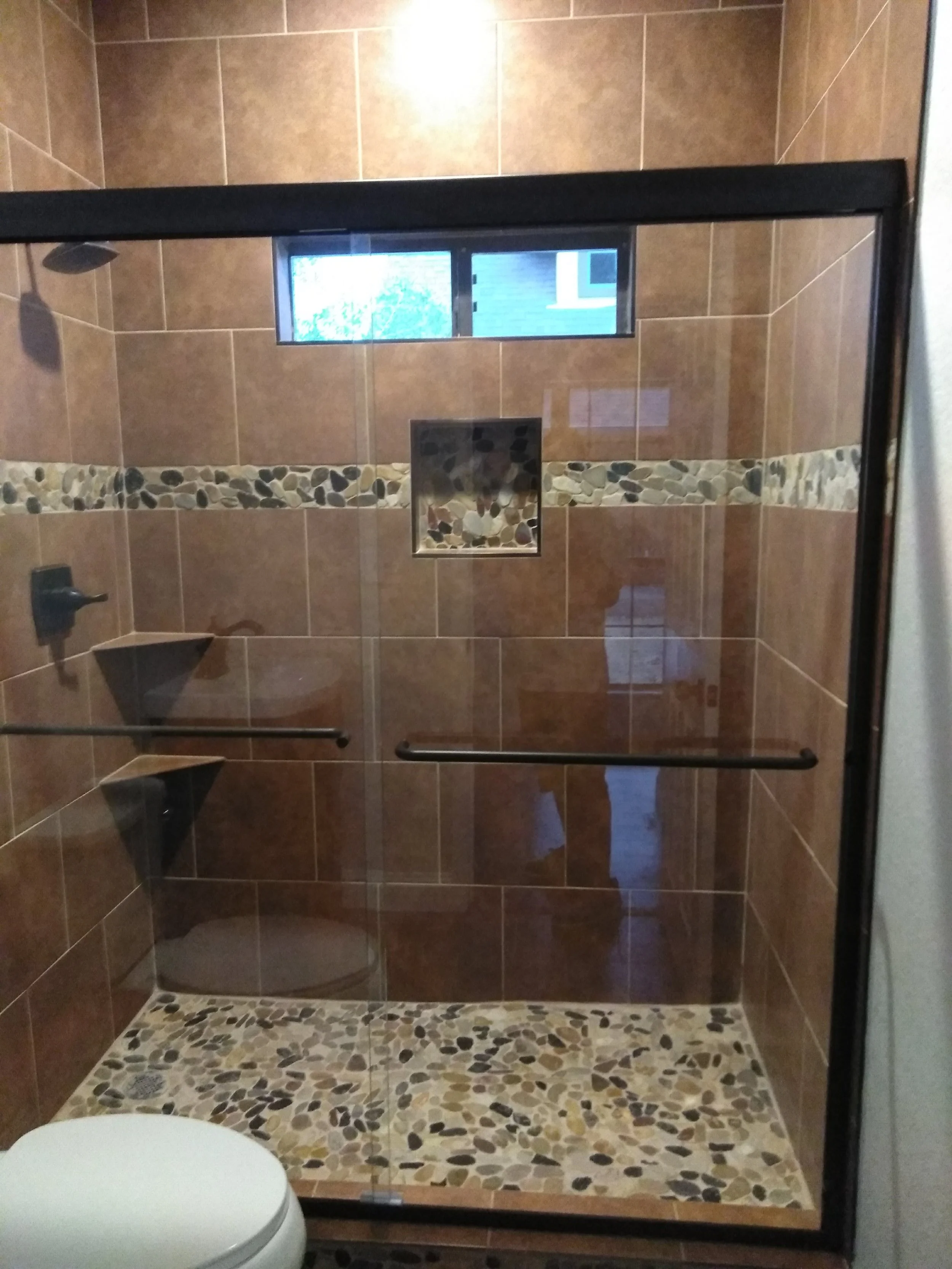 Shower with brown tiles, pebble accent strip, small window, and glass door, with a white toilet in the bathroom