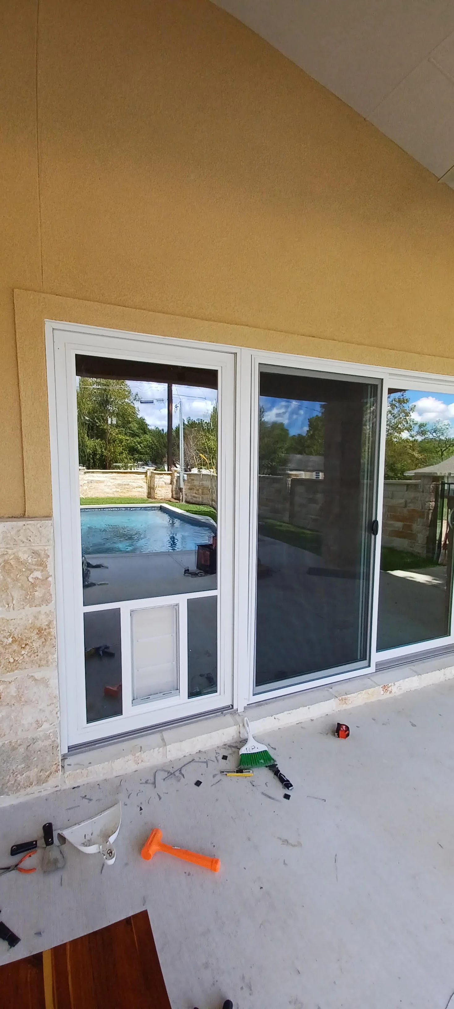 Looking through a sliding glass door onto a backyard with a swimming pool, stone wall, and green trees. The door frames and screen are visible inside, with construction tools and debris scattered on the floor.
