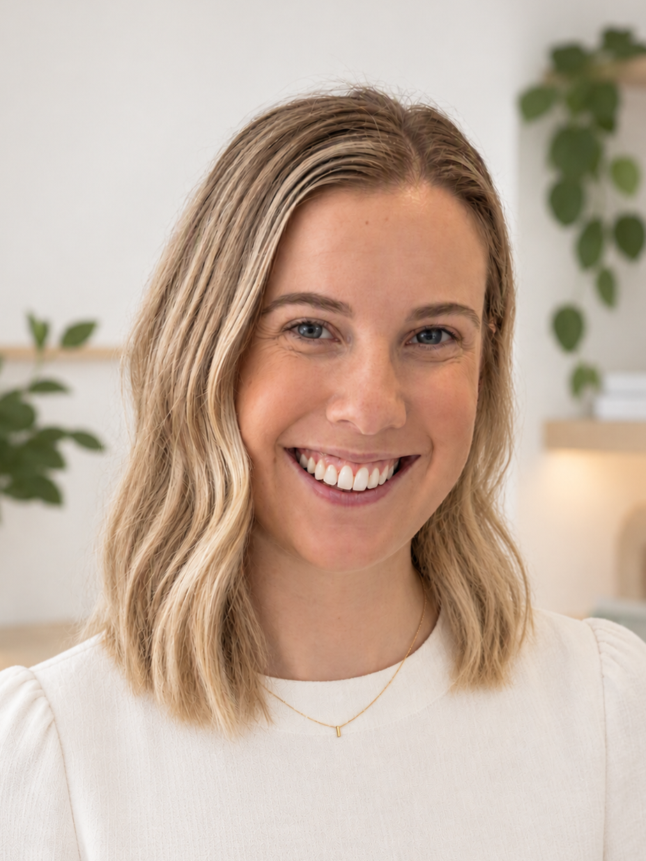 A smiling woman with shoulder-length blonde hair, blue eyes, and a white top standing indoors with a blurred background of green plants and shelves.