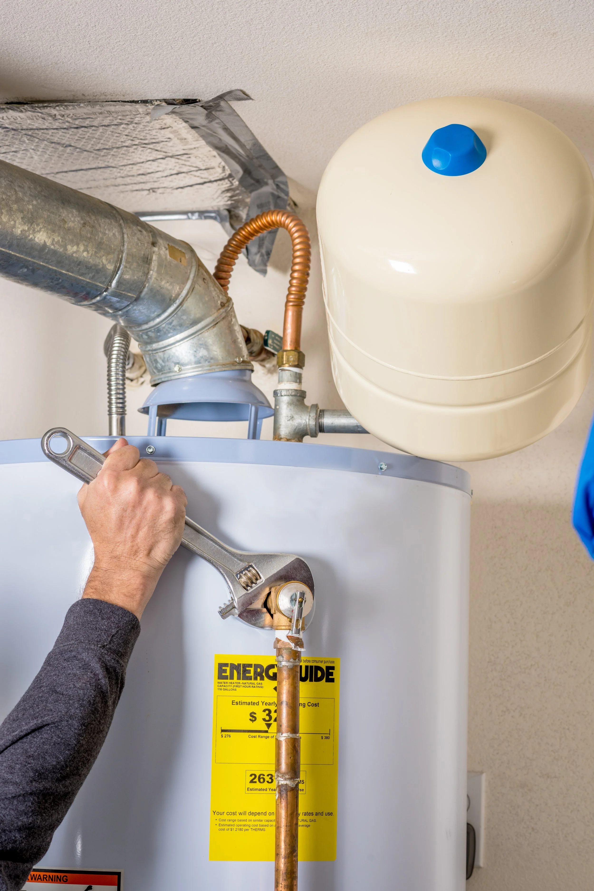 Person working on a water heater, adjusting a white panel with a stainless steel wrench, with pipes and a yellow energy guide label attached to the water heater.