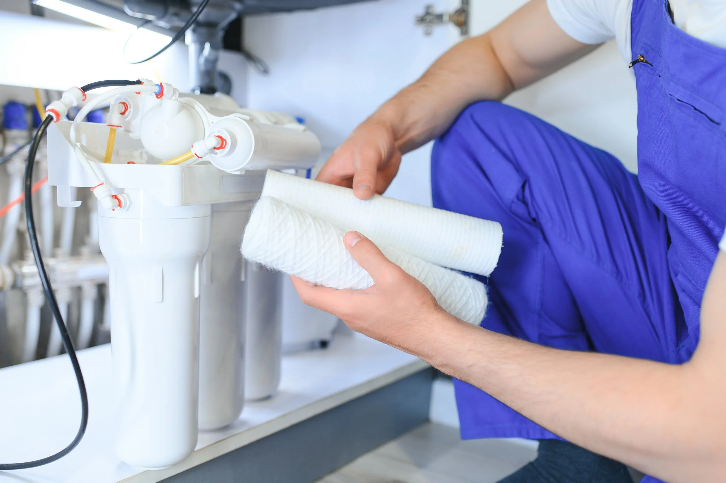 Person wearing blue scrubs repairing or installing a water filtration system under a sink, holding a white filter or membrane.