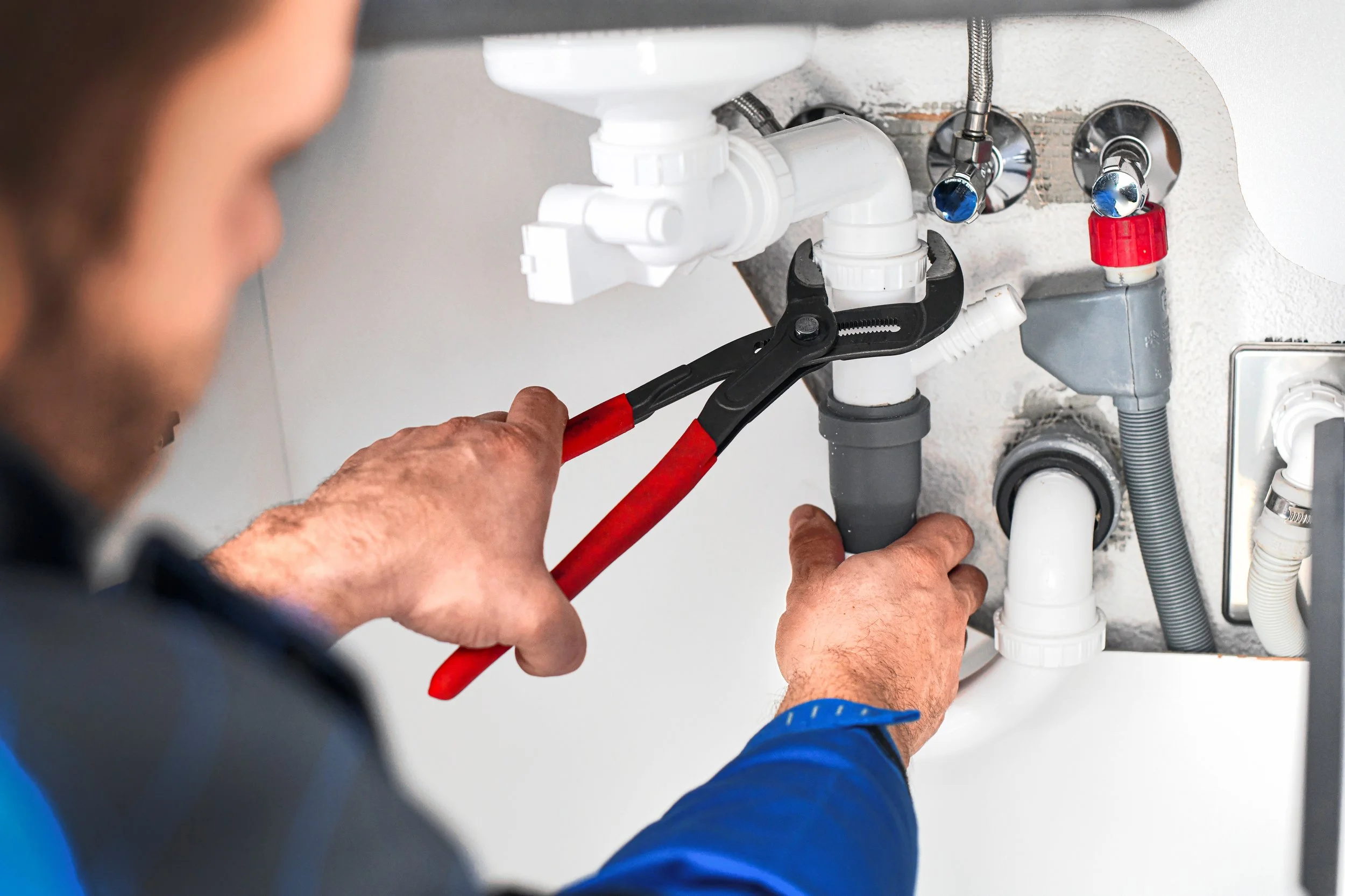 Person using pliers to work on plumbing pipes under a sink.