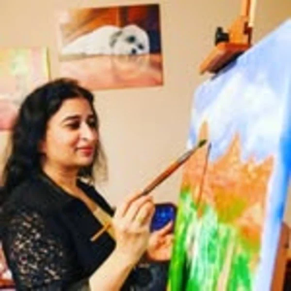 A woman with dark, curly hair wearing a black lace top is painting on a canvas set up on an easel. She is holding a paintbrush and a palette, working on a landscape scene with trees and sky. There is a picture of a dog on the wall behind her.