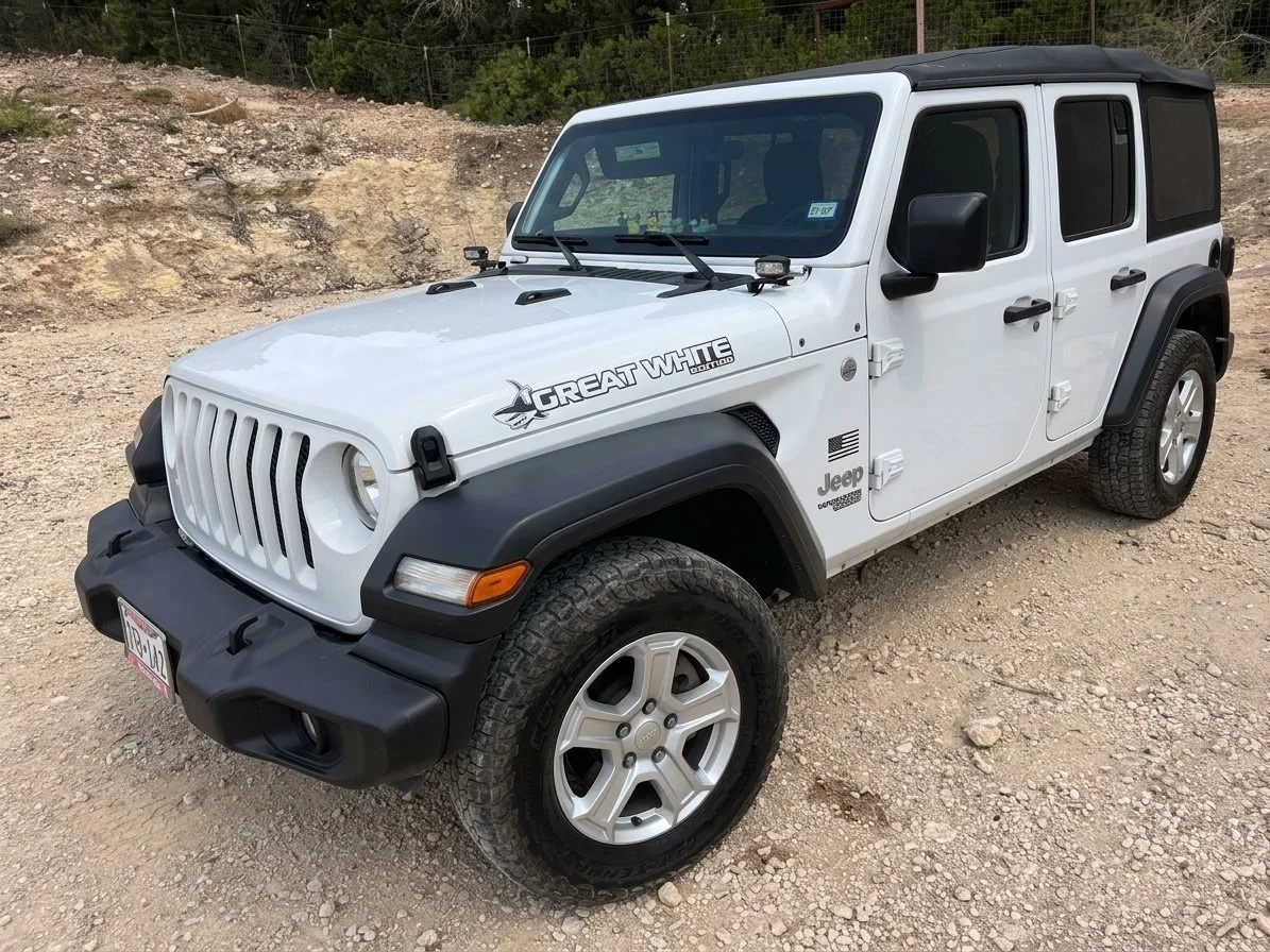 White Jeep Wrangler with black accents parked on dirt ground near a rocky hill and trees, with decals reading 'Great White' and 'Jeep' on the side.