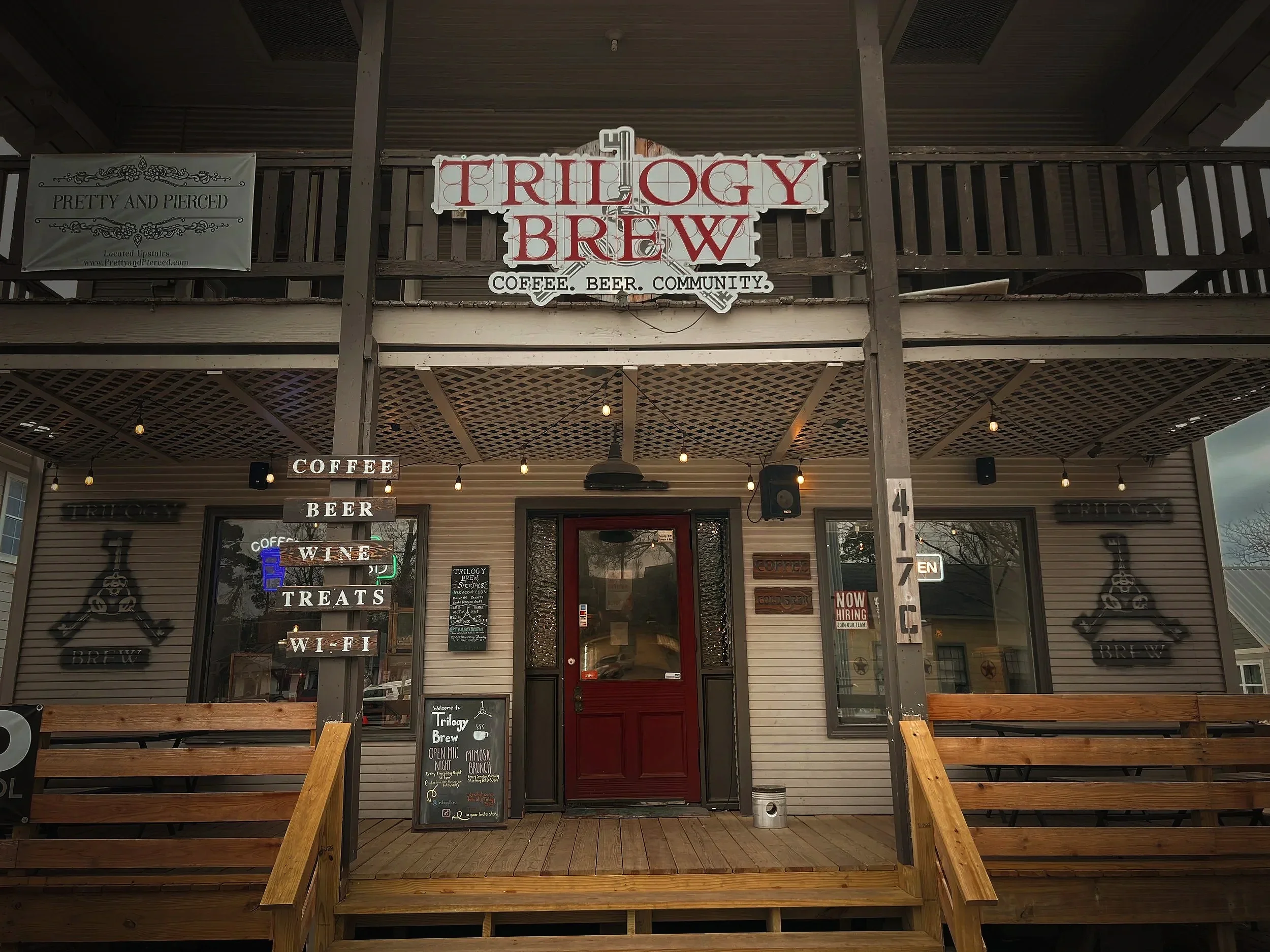 Exterior of Trilogy Brew coffee and beer venue with wooden patio seating, string lights, and signs for coffee, beer, wine, treats, and community.
