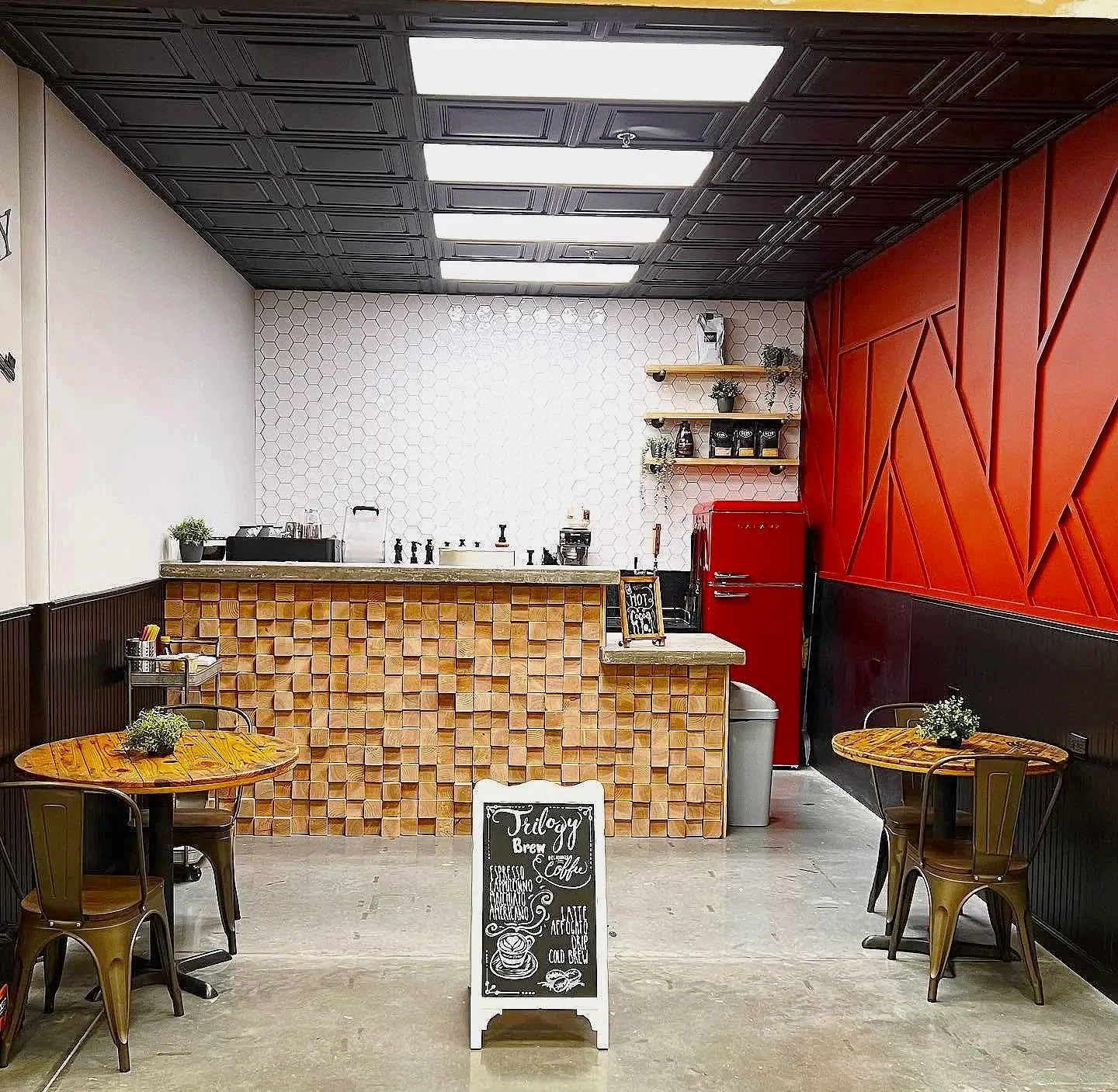 Stylish coffee shop interior with a wood-tile counter, hexagonal backsplash, red refrigerator, café tables, and chalkboard menu.