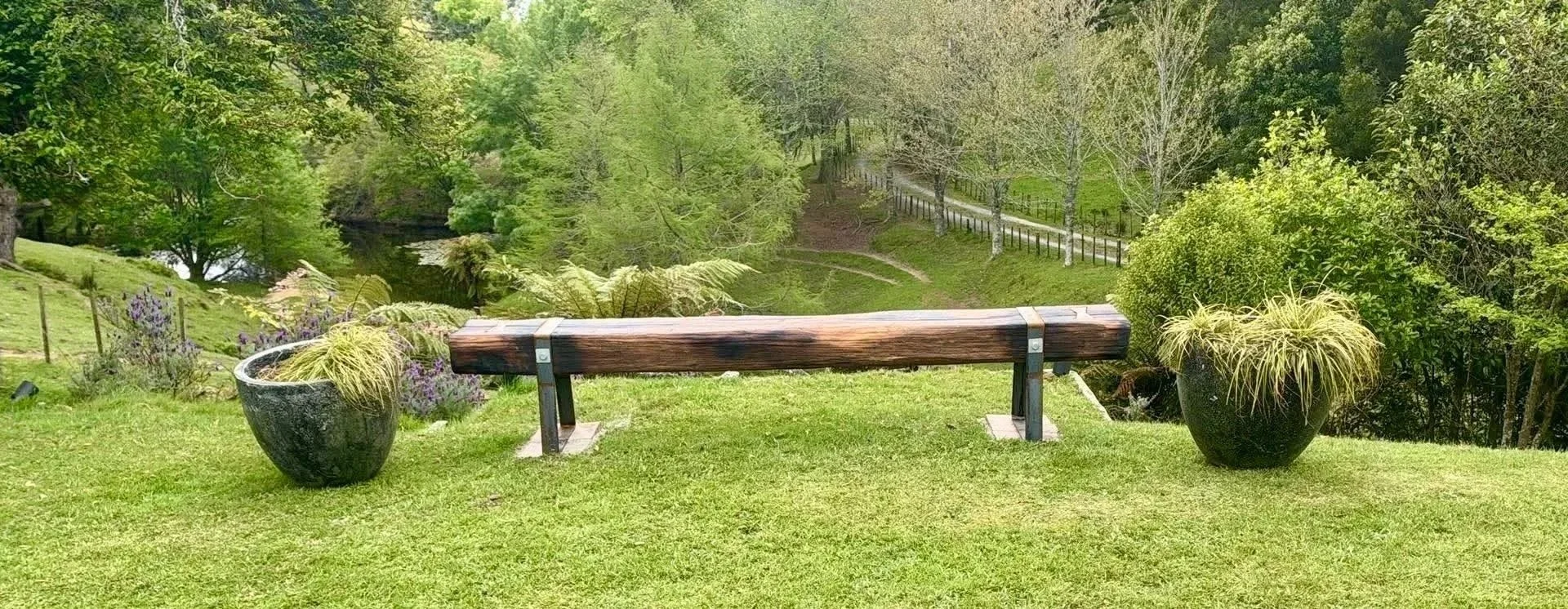 Designer solid timber outdoor bench with steel legs, positioned in a landscaped New Zealand garden surrounded by greenery.