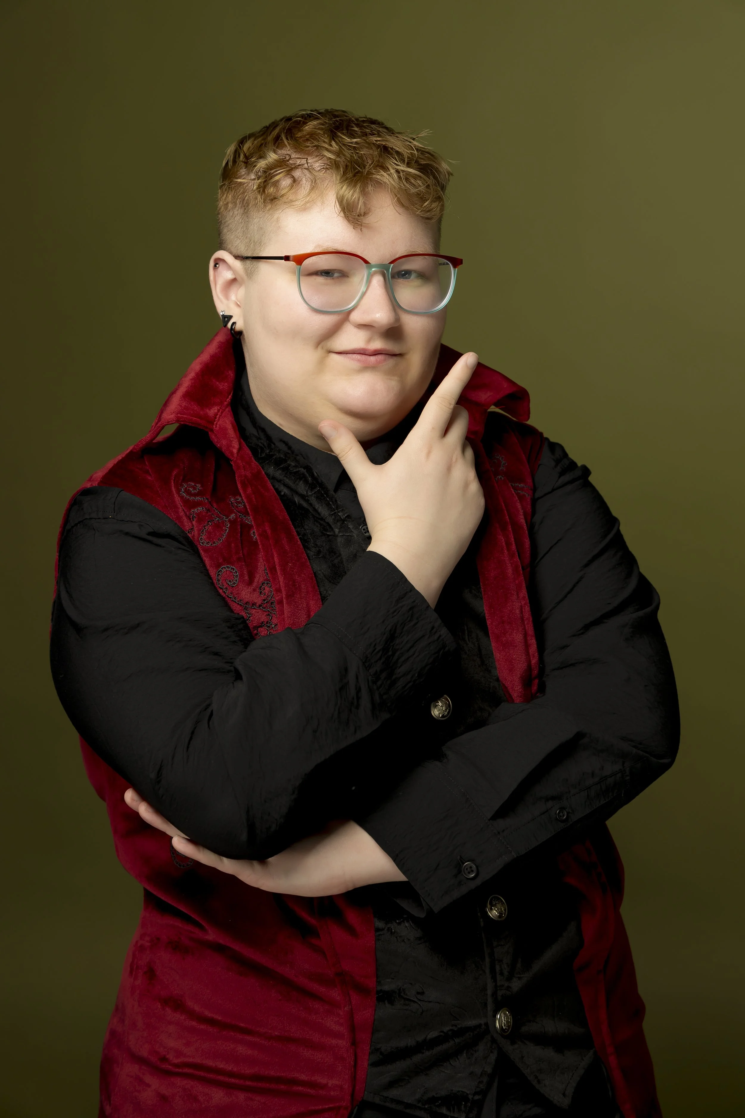 Person wearing multicolored glasses and a red vest over black clothing, posing with arms crossed against an olive-green background