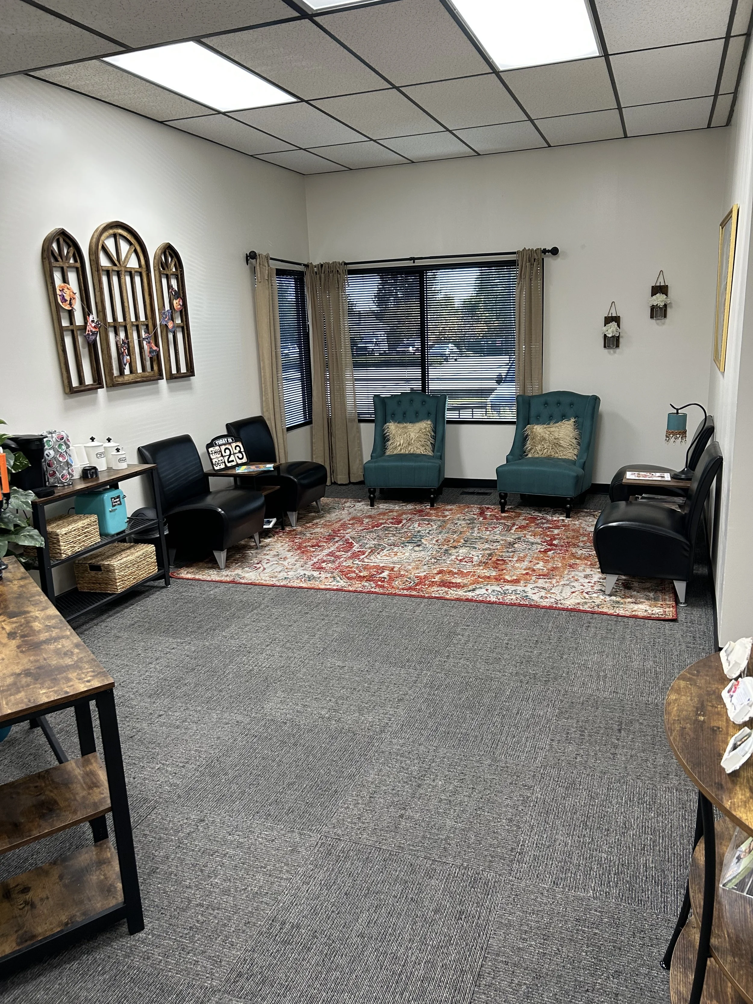 Cozy office waiting room with teal armchairs, black leather chairs, patterned area rug, windows with blinds, and decorative wall shelving.