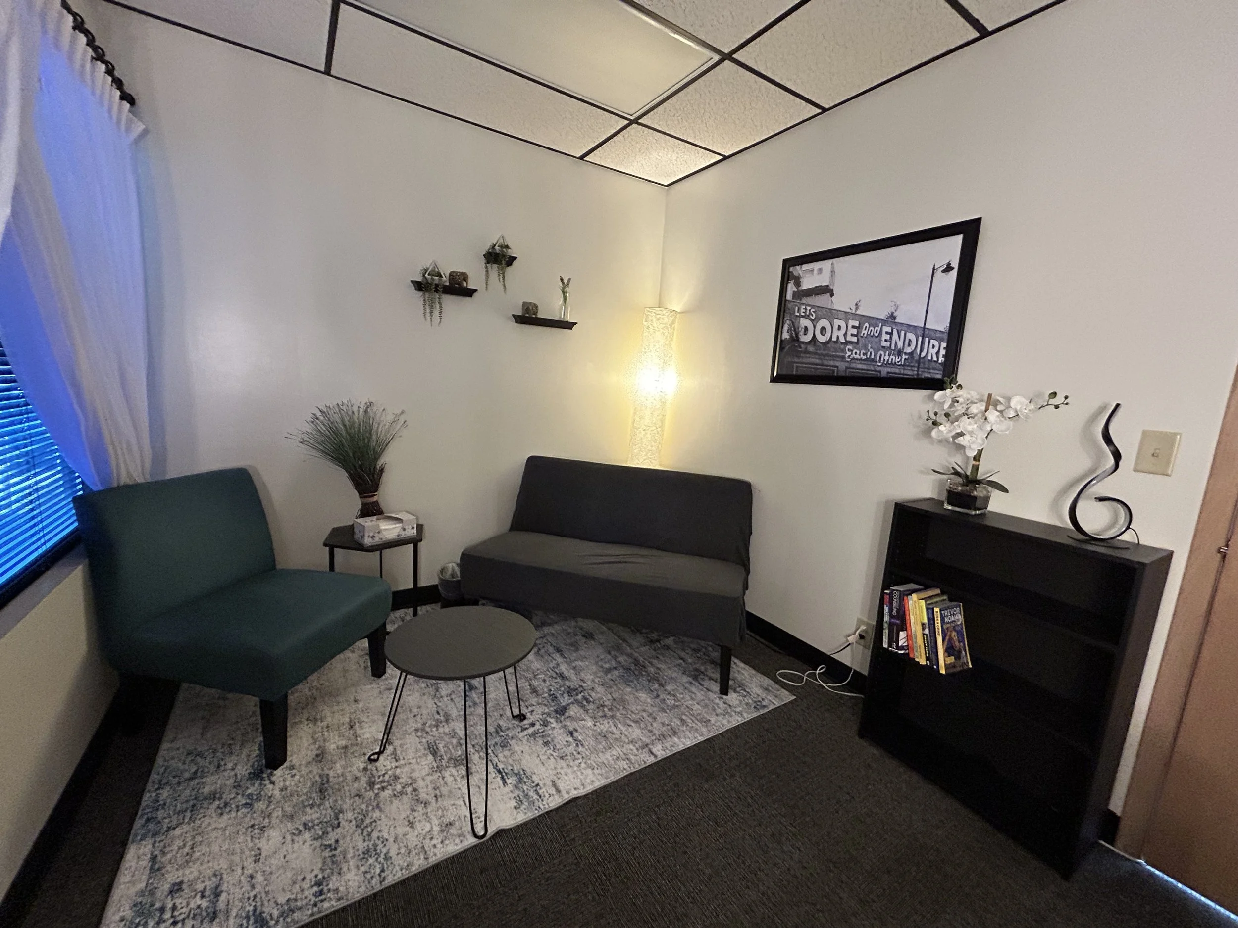 Cozy modern office lounge with a gray loveseat, teal armchair, round coffee table, floor lamp, decorative shelves, and black bookcase.