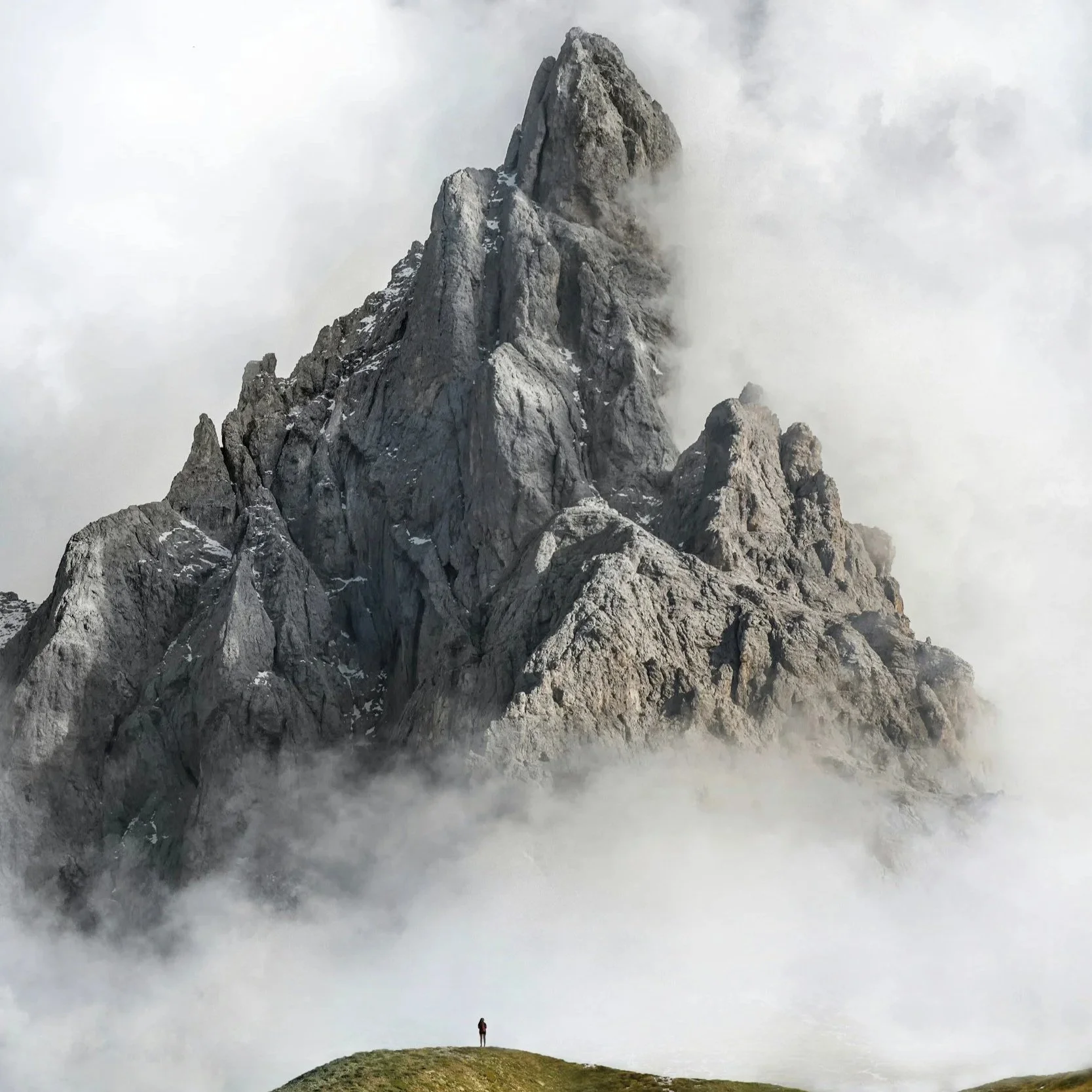 A ginormous, rocky mountain, half covered with fog, with a person standing at it's base and looking up, making them seem tiny. tiny in comparison